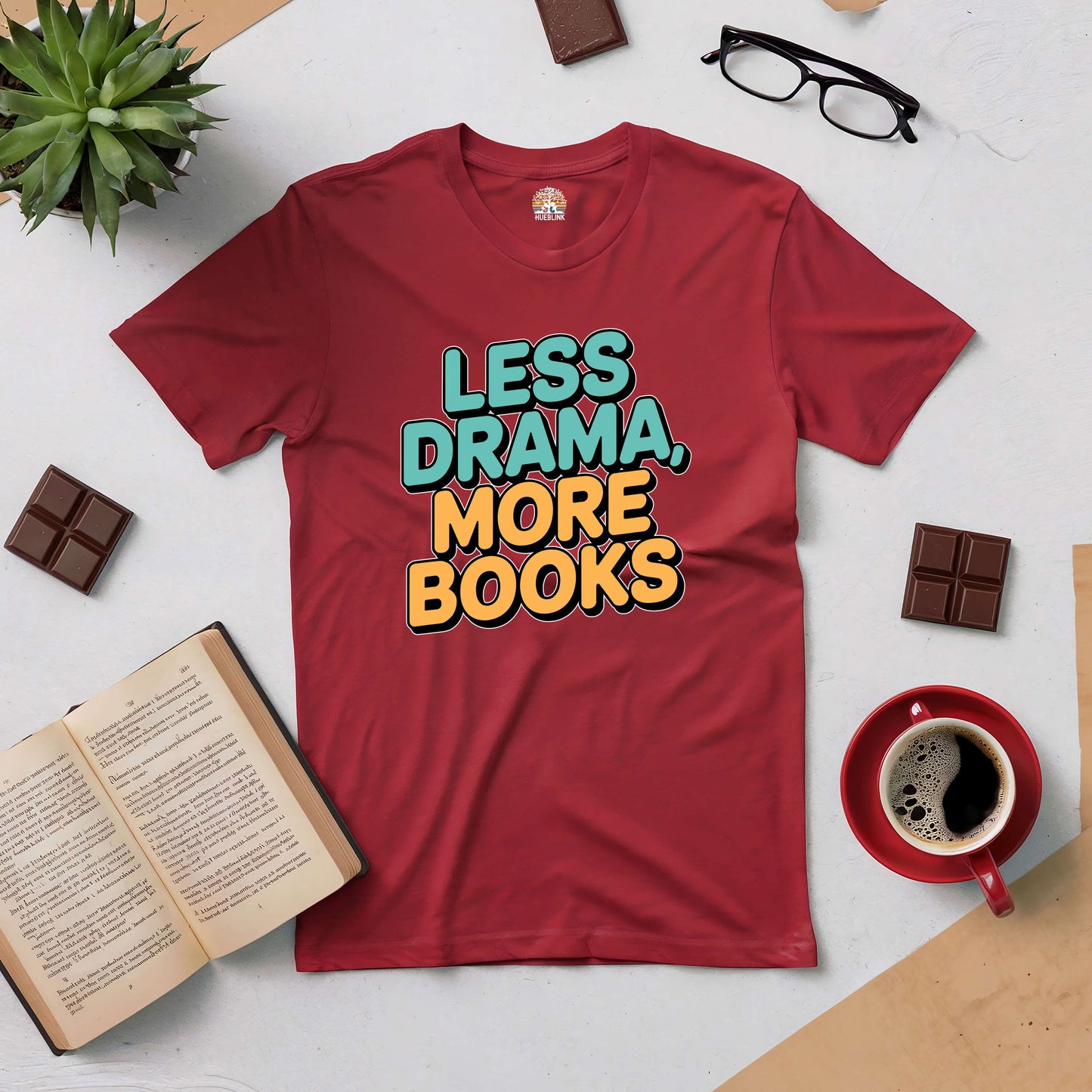 Red 'Less Drama, More Books' tee surrounded by a book, coffee, glasses, chocolate, and a plant, perfect for book lovers.