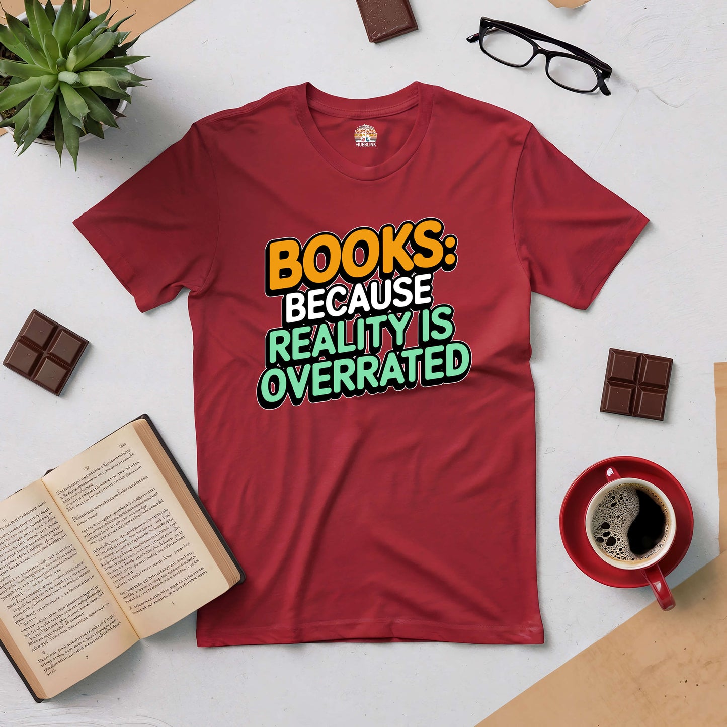 Red t-shirt with text "Books: Because Reality Is Overrated," surrounded by a book, glasses, coffee, chocolate, and a plant.