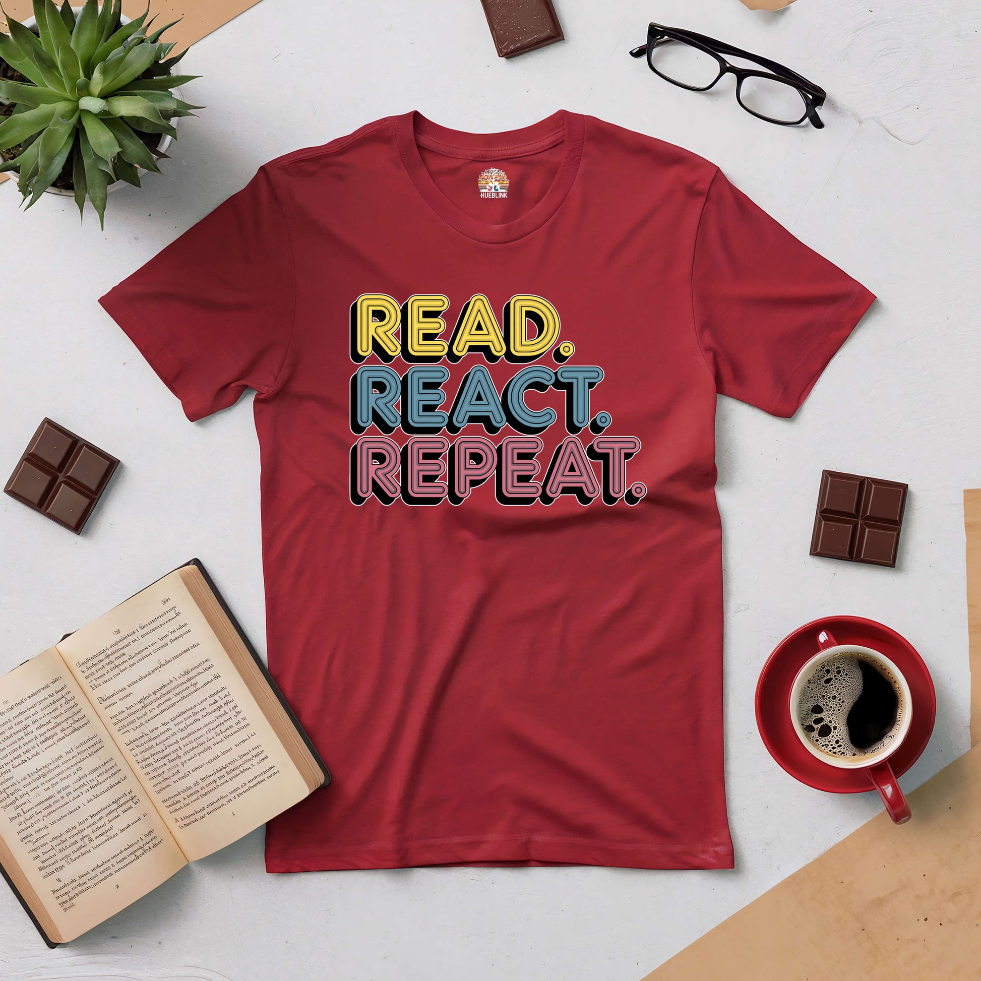 Red "Read. React. Repeat." tee with open book, coffee, glasses, chocolate, and plant on a light background for book lovers.