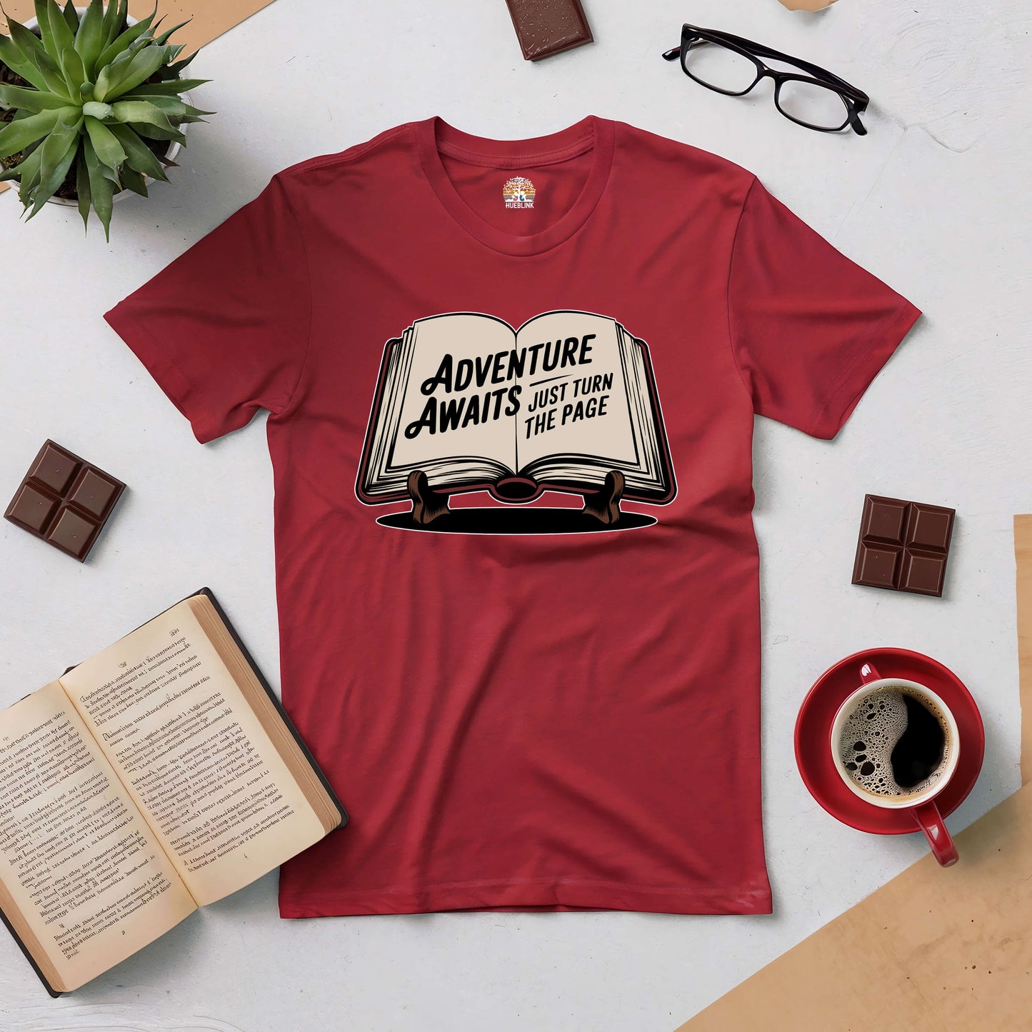 Red 'Adventure Awaits: Just Turn the Page' tee surrounded by books, glasses, coffee, and chocolate on a white background.