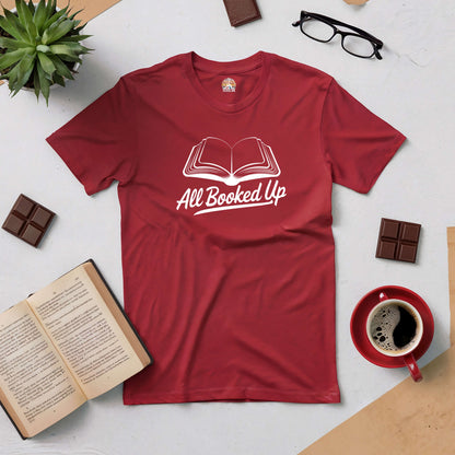 Red All Booked Up Tee with open book design, surrounded by coffee, chocolate, glasses, and a plant, perfect for avid readers.