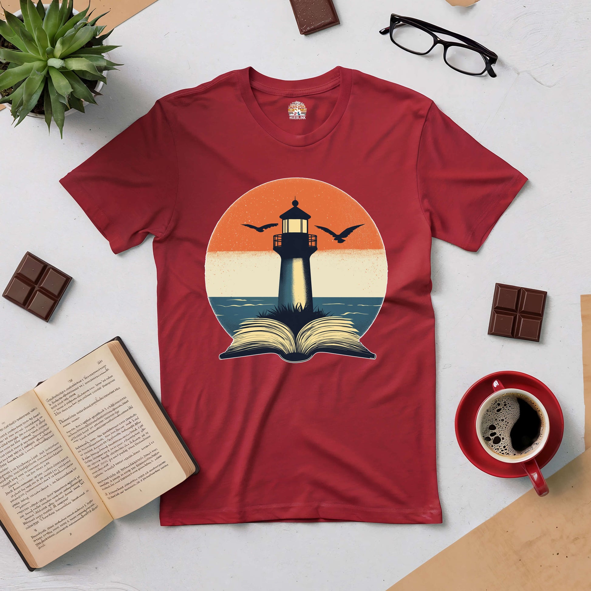 Red tee with a lighthouse and open book design, symbolizing guidance and exploration. Surrounded by coffee, chocolate, and reading glasses.