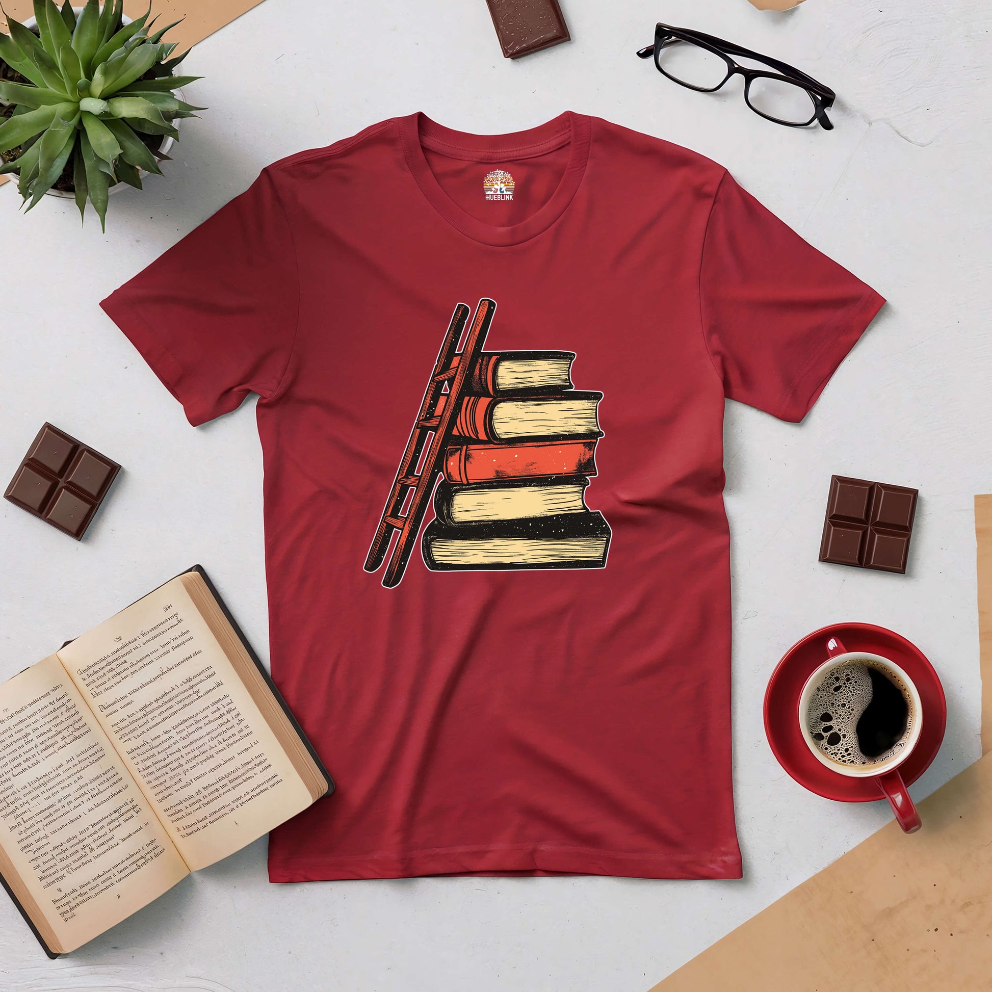 "Reaching New Heights Tee with ladder and book design, symbolizing readers' journey of knowledge, displayed with coffee and books"