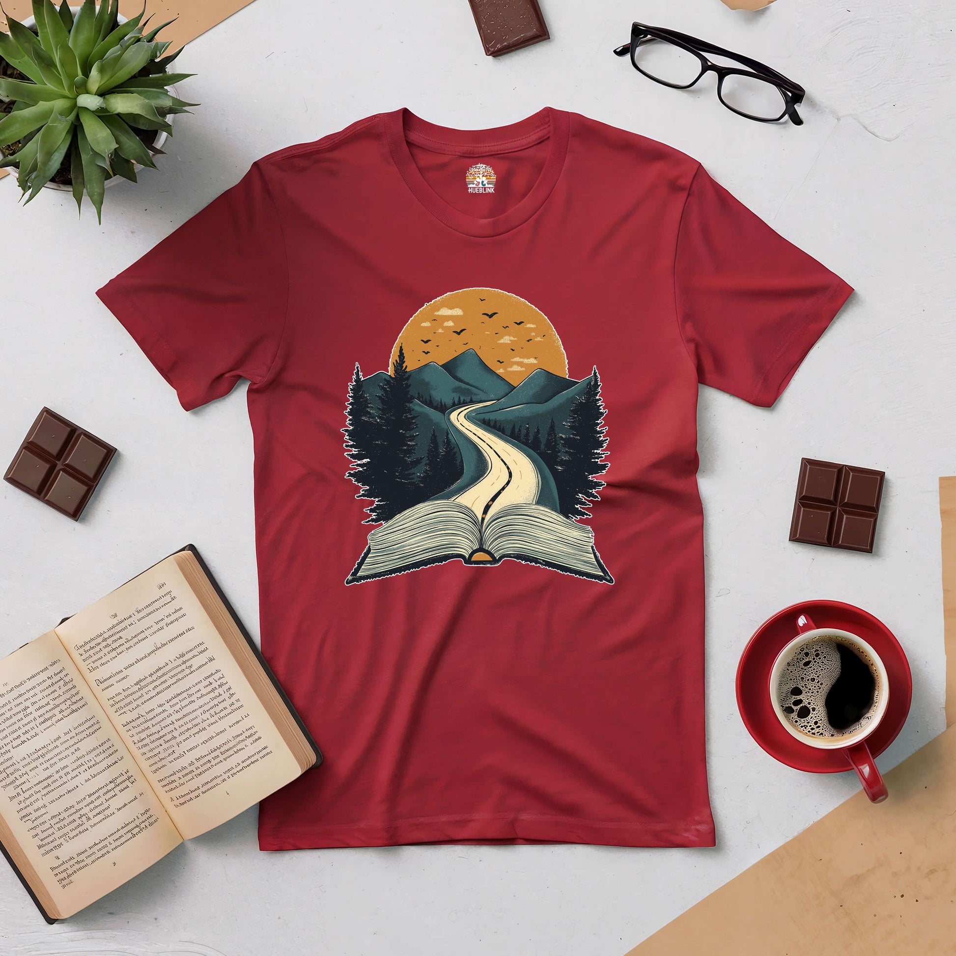 Red Starting Point Tee with book and mountain design, surrounded by coffee, an open book, and chocolate, ideal for readers and adventurers.