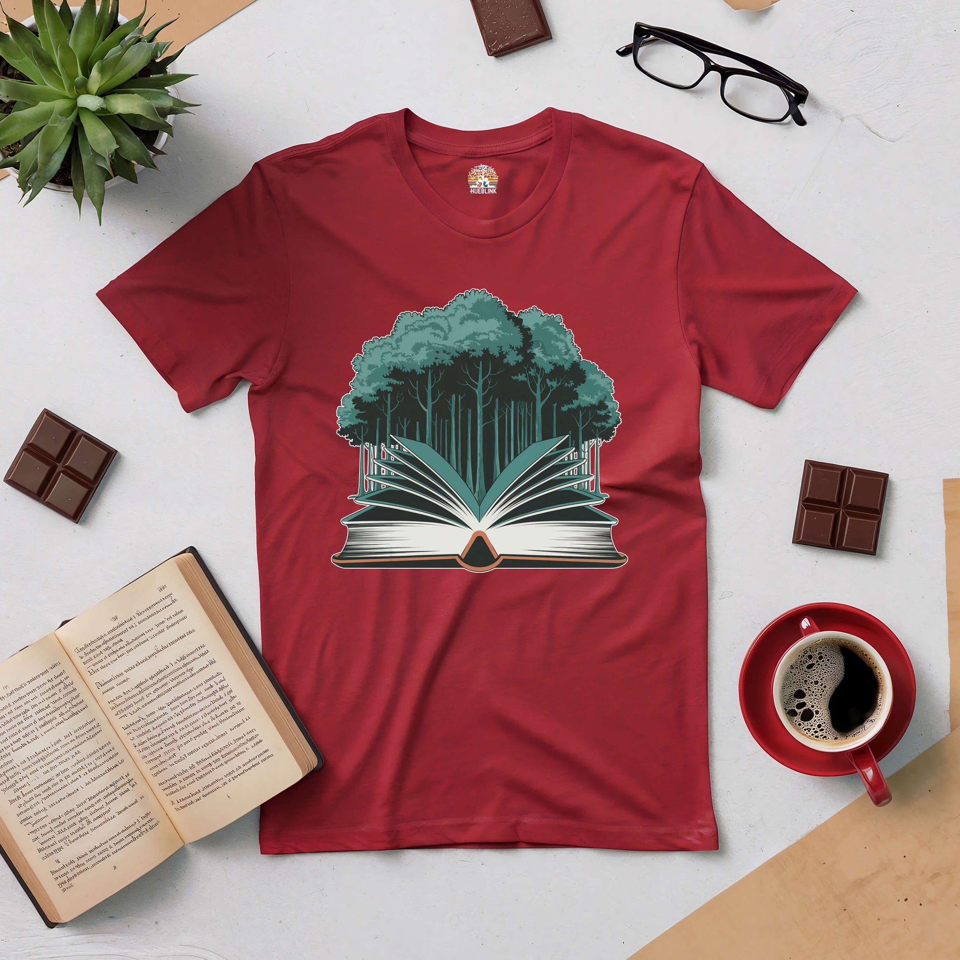 Red "Rooted in Story" tee with book and forest design, surrounded by coffee, chocolate, glasses, and open book on a table.