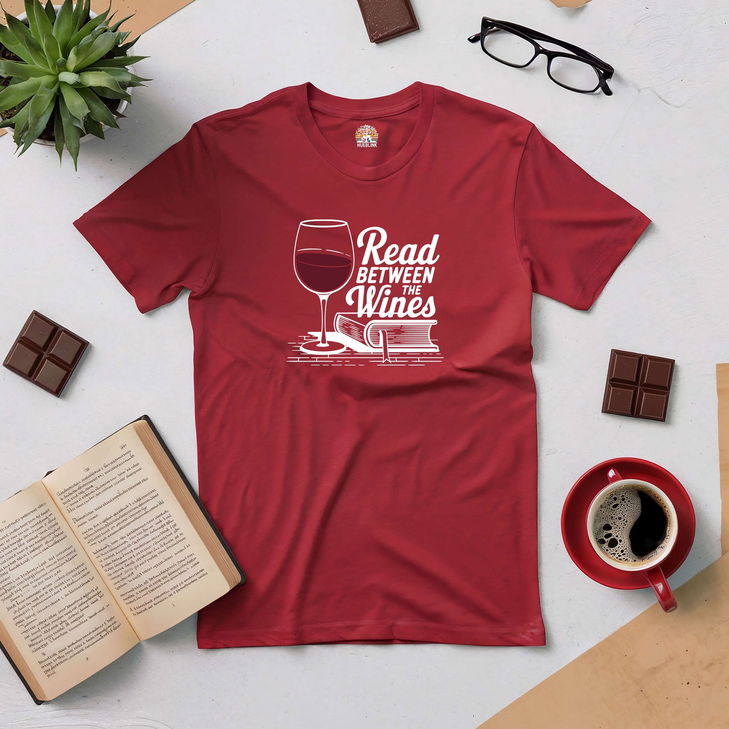 Red "Read Between the Wines" tee with book and wine glass design, surrounded by coffee, glasses, chocolate, and an open book.