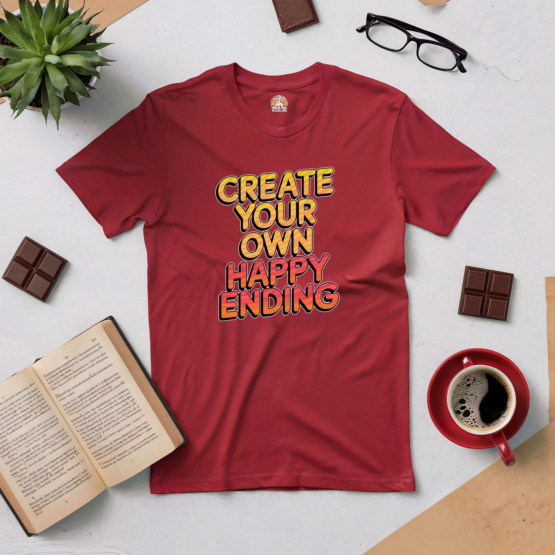 Red tee with "Create Your Own Happy Ending" slogan, surrounded by coffee, book, glasses, plant, and chocolate bars on a table.