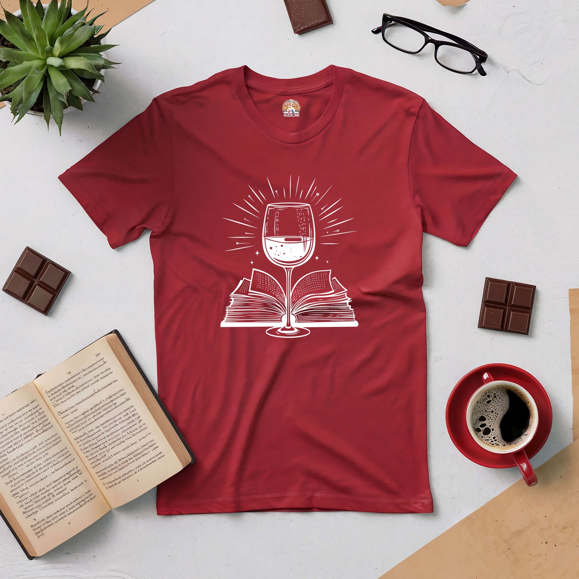 Red Literary Libations tee with wine glass and book design, surrounded by chocolate, coffee, and open book on a flat lay surface.