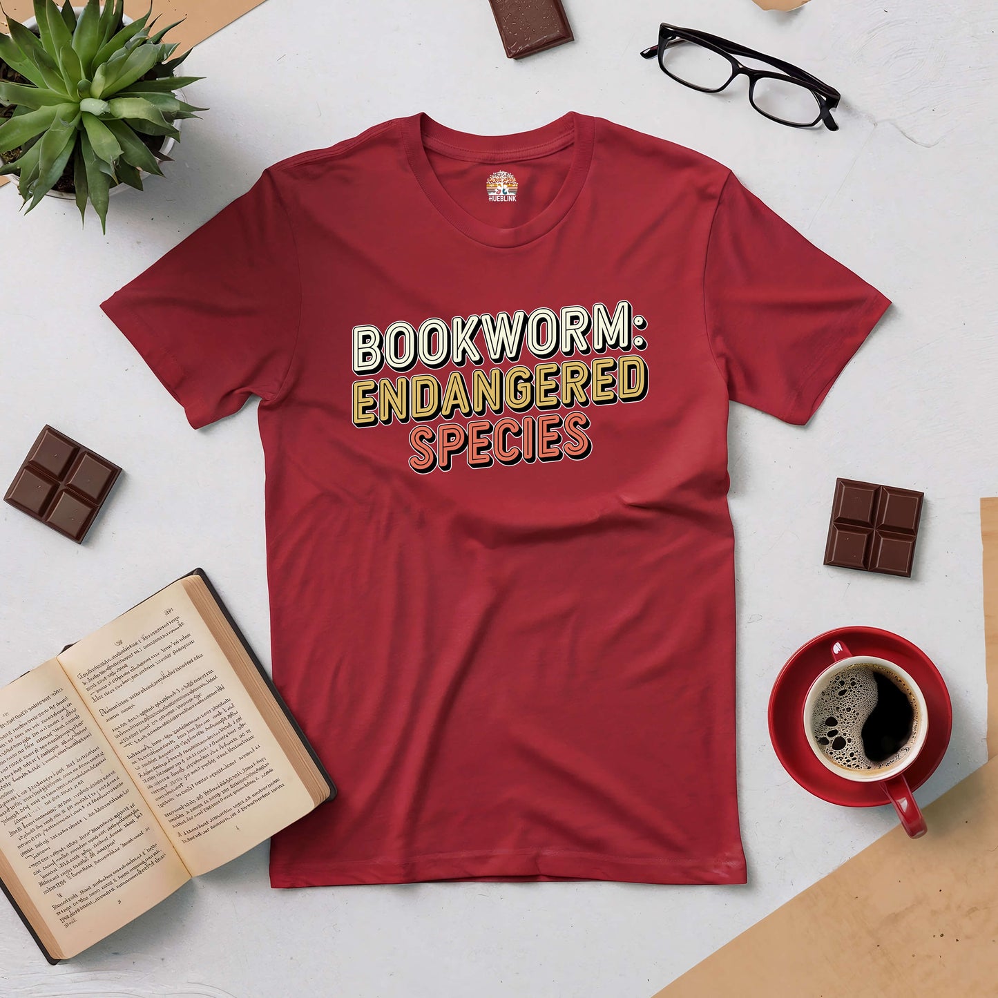 Red "Bookworm: Endangered Species" tee surrounded by books, coffee, glasses, and chocolate squares, perfect for book lovers.