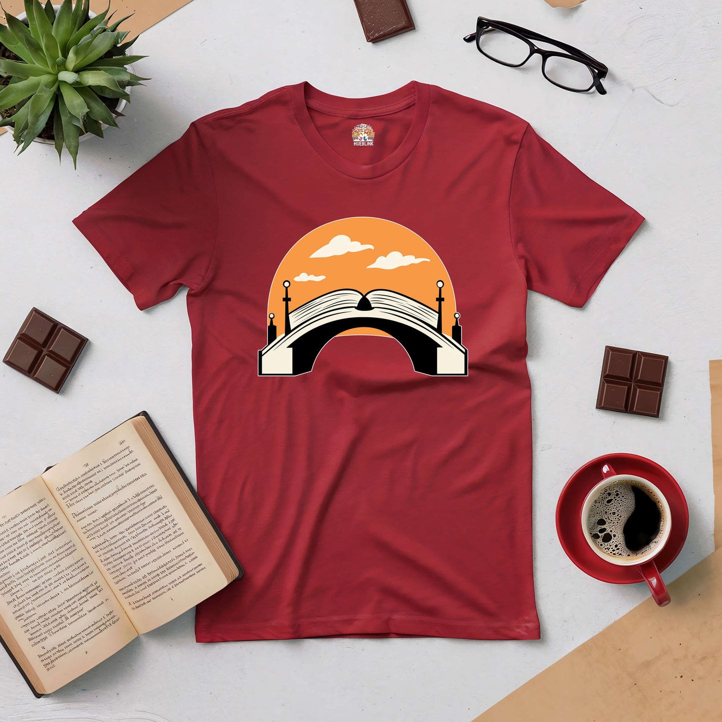 Red "Connection Through Decades" tee featuring a bridge and open book design amidst coffee, chocolate, and reading items.