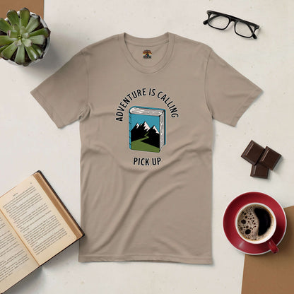 Adventure Is Calling Pick Up Tee with book graphic, surrounded by coffee, chocolate, and glasses on a cozy desk setup.