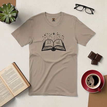 "Stellar Script Tee with open book and stars design on beige shirt, surrounded by coffee, chocolate, glasses, and a plant."