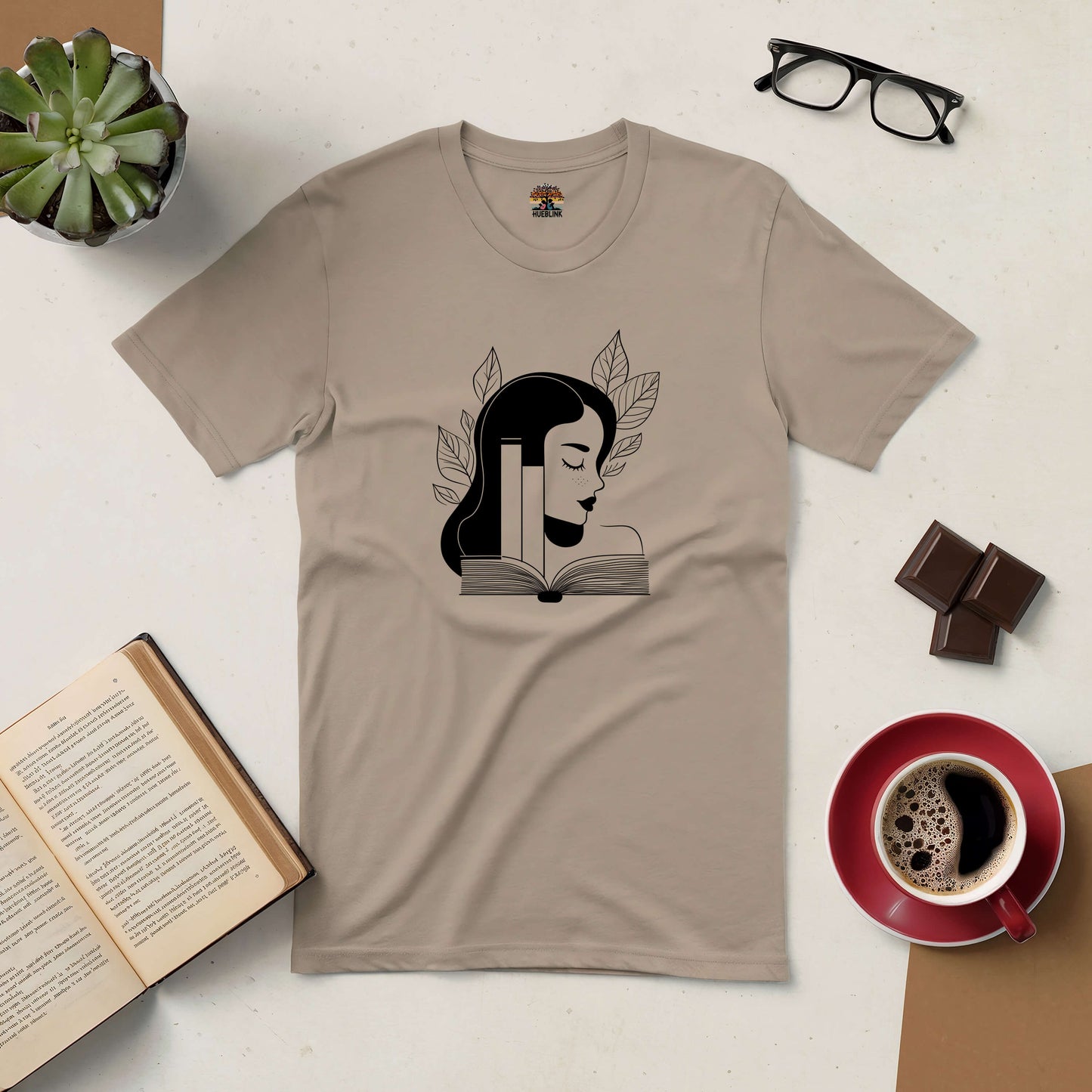 Beige "Beauty in Every Page" tee with artistic illustration of a woman and books, styled with open book, coffee, and glasses.