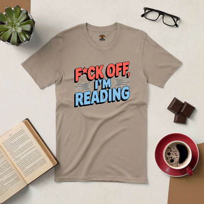 Beige tee with "F*ck Off, I'm Reading" text, surrounded by coffee cup, book, glasses, chocolate, and plant on table.