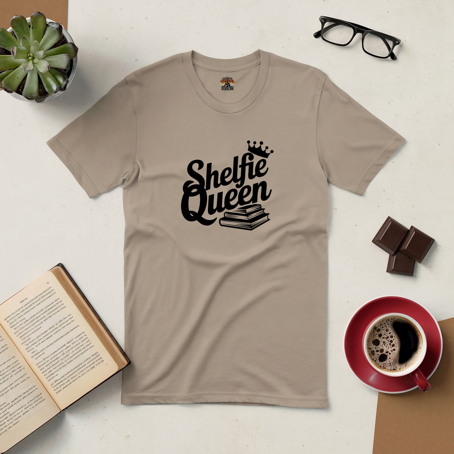 "Shelfie Queen Tee for book lovers with coffee and open book, featuring black text and crown design on beige shirt."