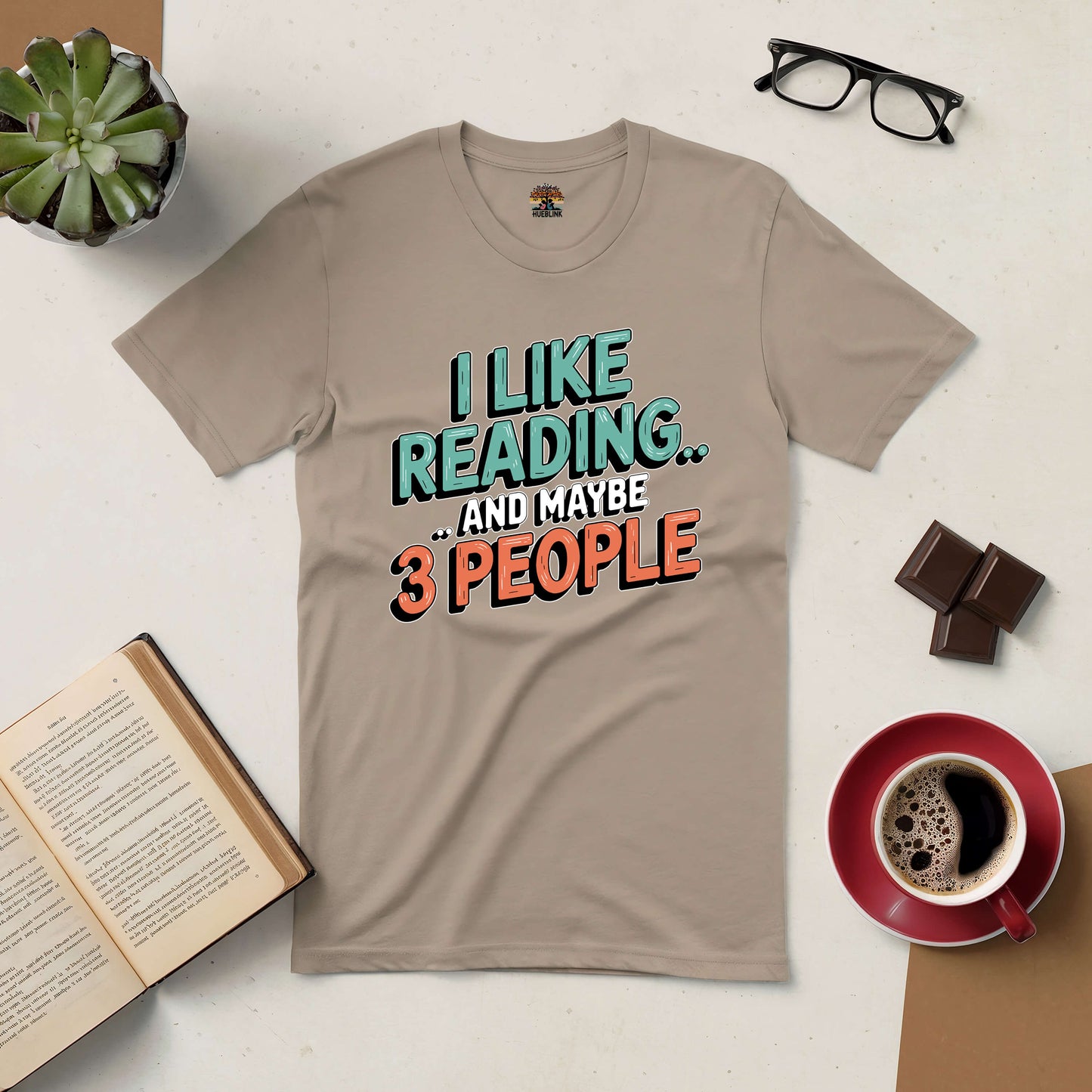 Beige tee with "I Like Reading... and Maybe 3 People" text, surrounded by books, coffee, glasses, and chocolate. Perfect for book lovers.