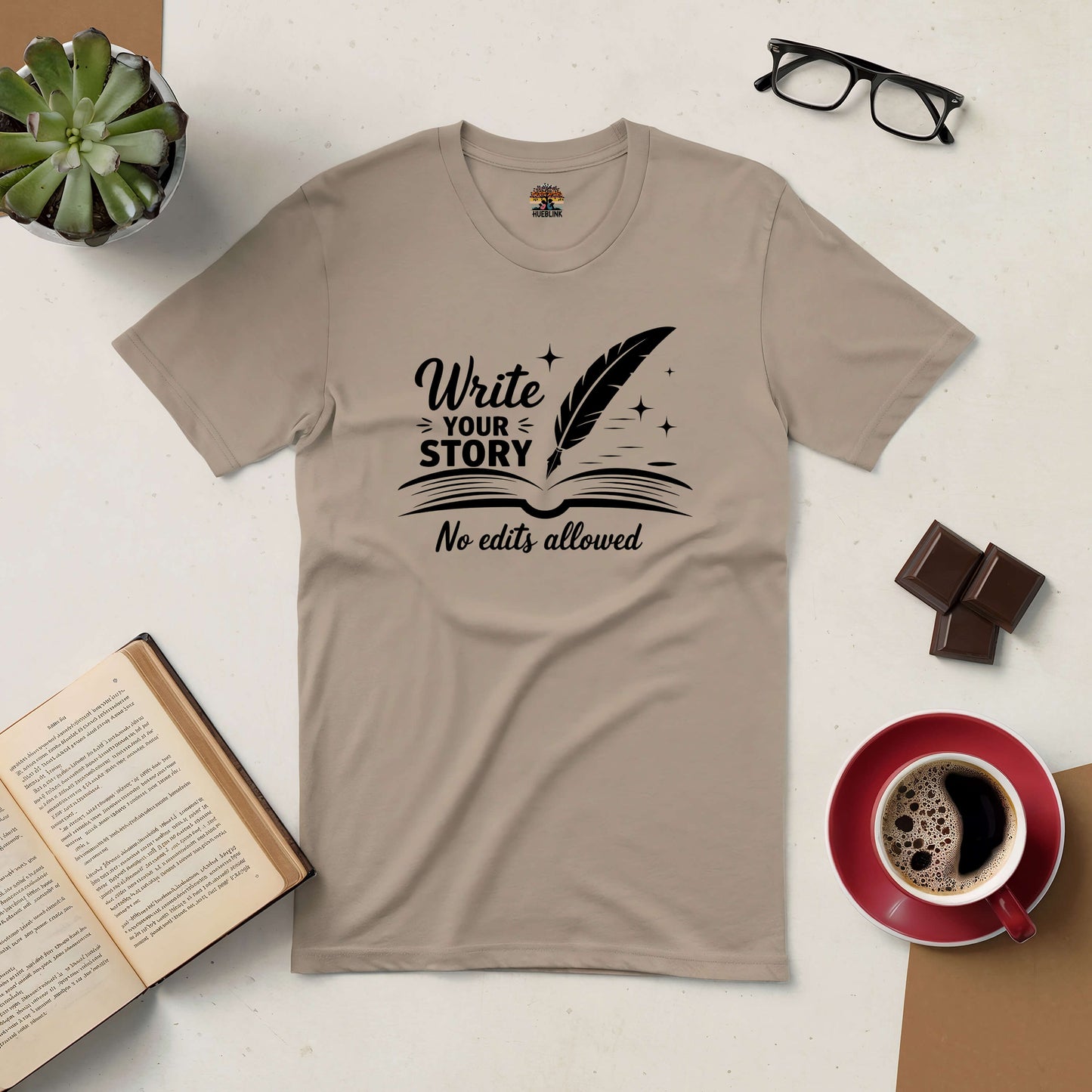 Beige t-shirt with "Write Your Story, No Edits Allowed" print, surrounded by coffee, glasses, a plant, chocolate, and an open book.