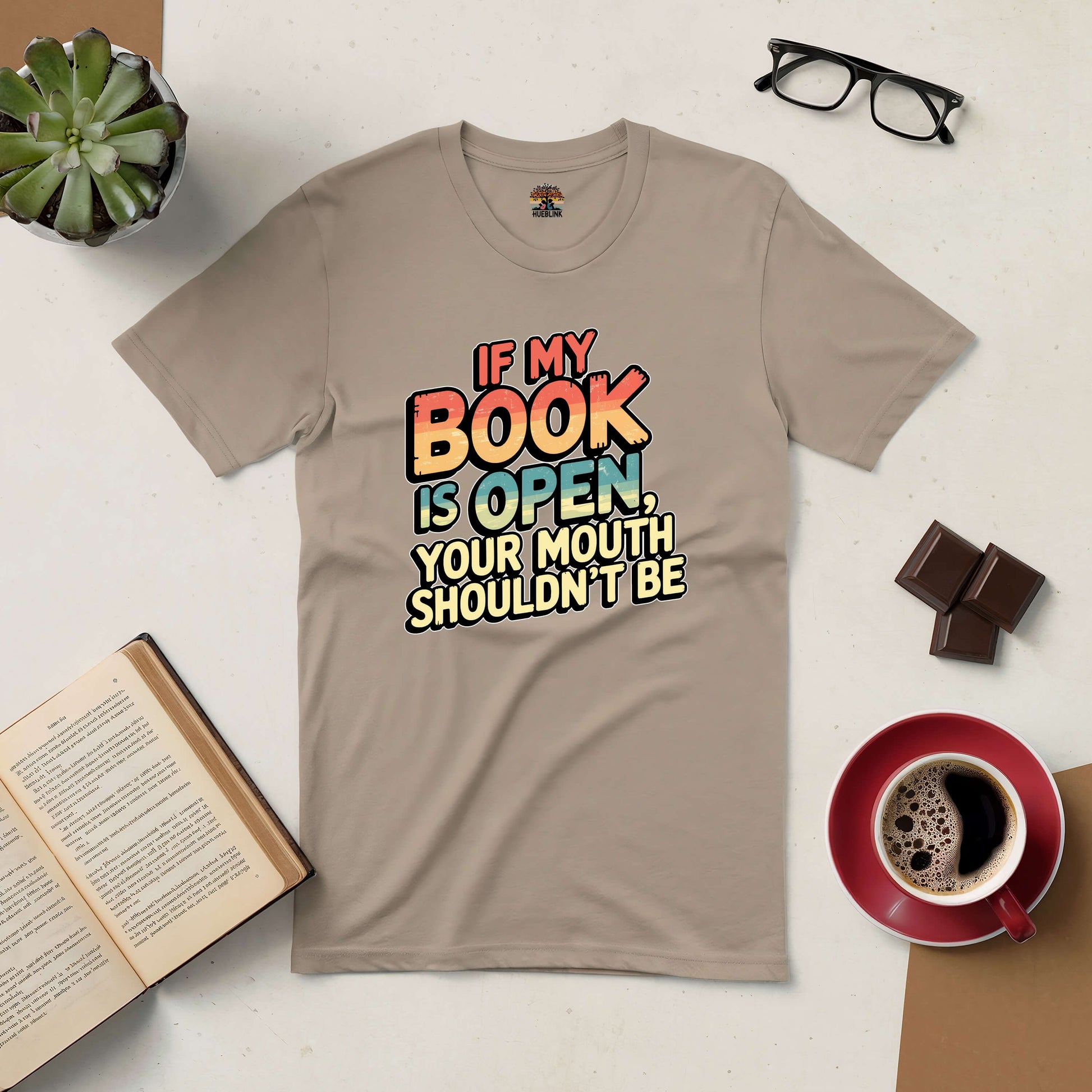 Beige tee with text "If My Book Is Open, Your Mouth Shouldn't Be" next to coffee, book, glasses, plant, and chocolate. Perfect for book lovers.