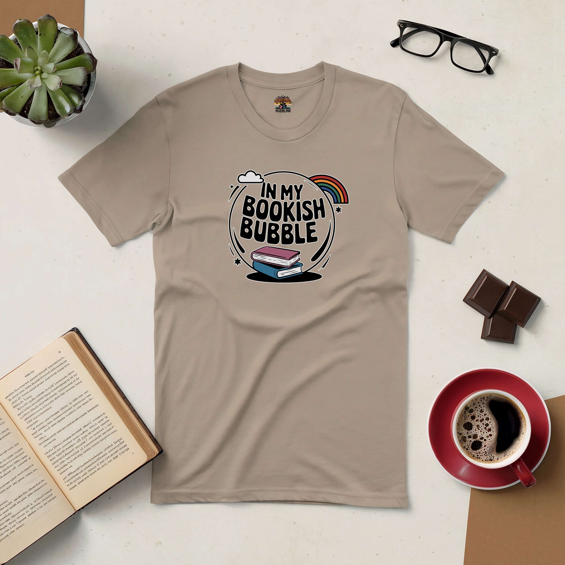 Beige tee with "In My Bookish Bubble" design featuring books, surrounded by coffee, glasses, chocolate, plant, and open book.