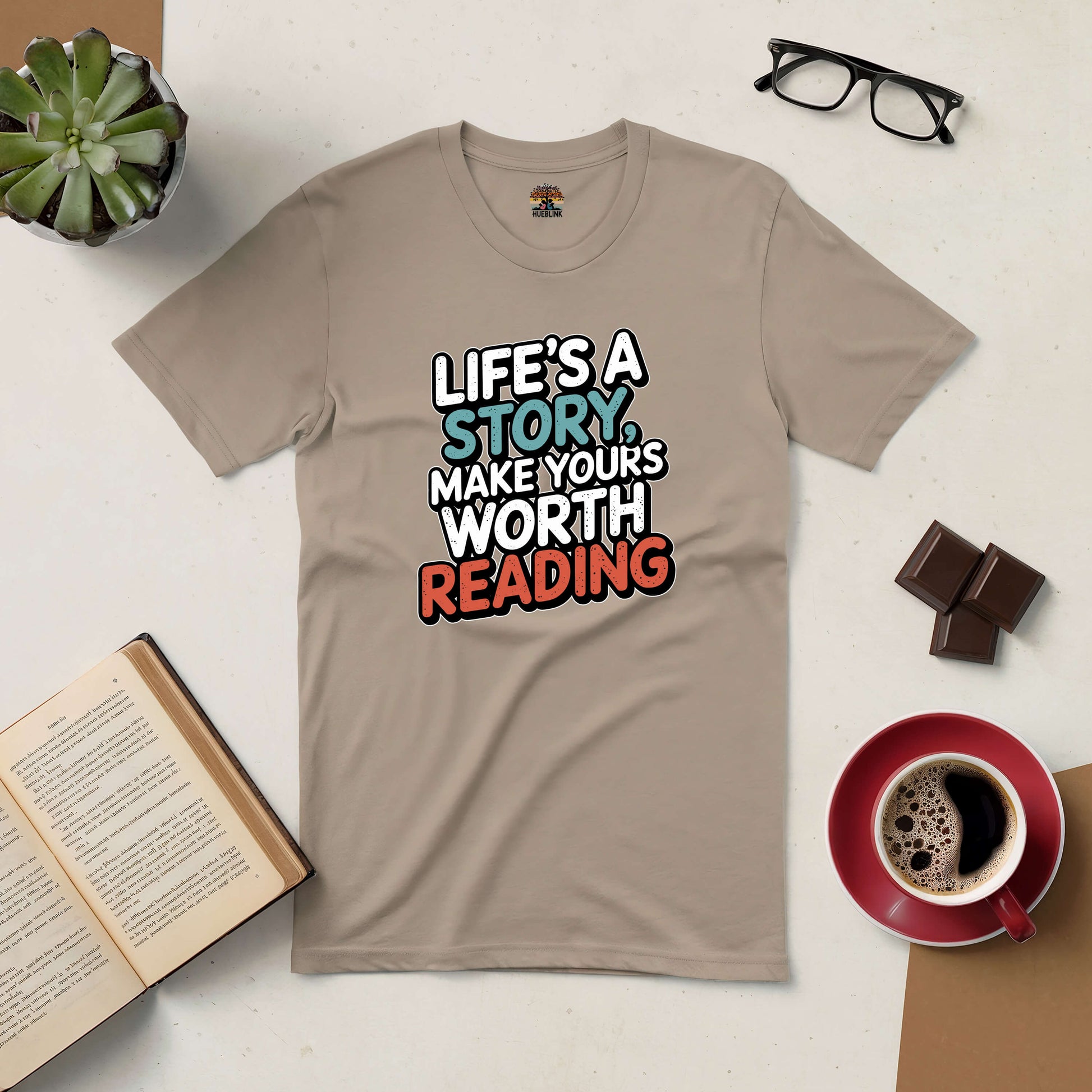 "Life's a Story Make Yours Worth Reading Tee with motivational text, surrounded by glasses, coffee, book, chocolate, and plant."