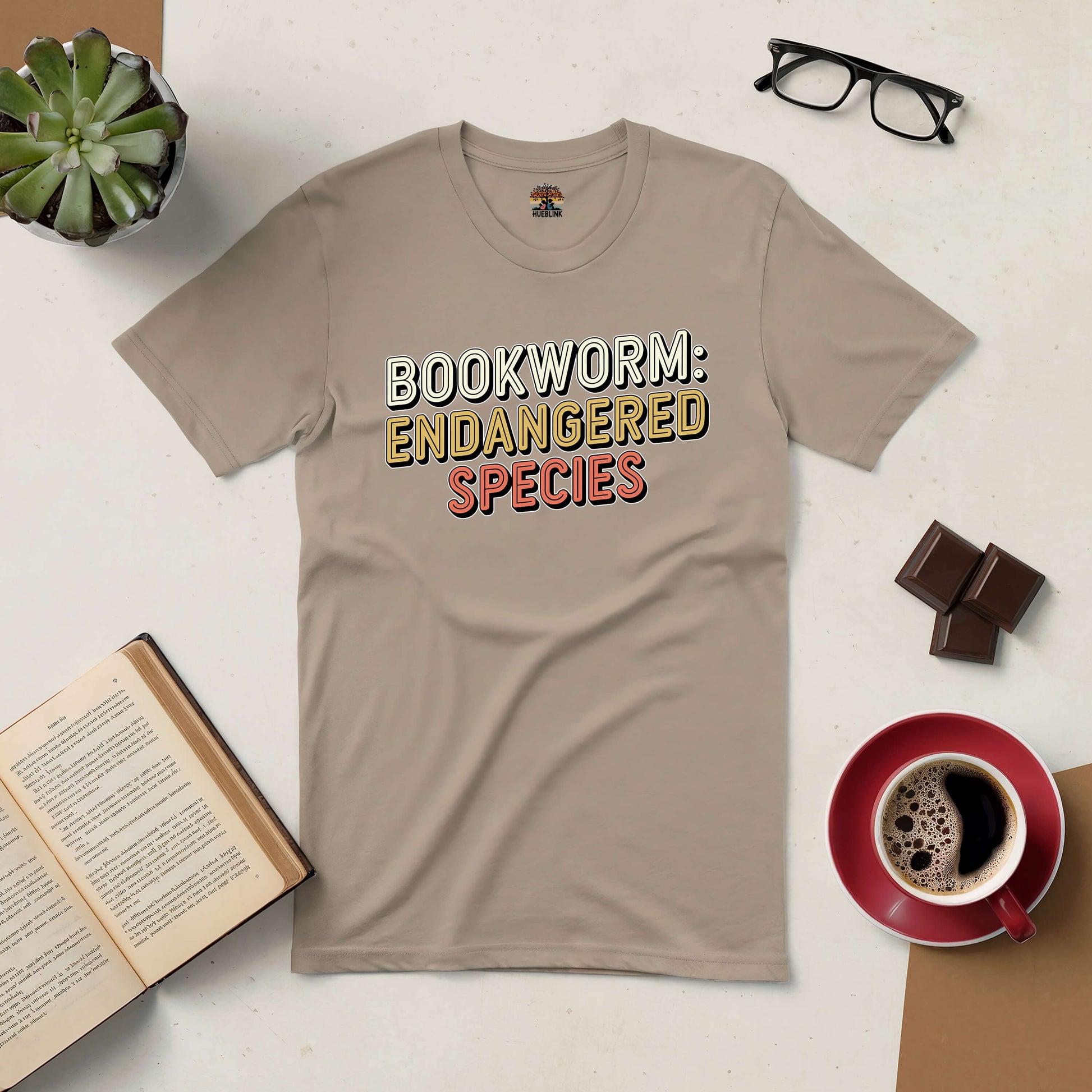 Beige "Bookworm: Endangered Species" tee with books, coffee, glasses, and chocolate, ideal for book lovers.