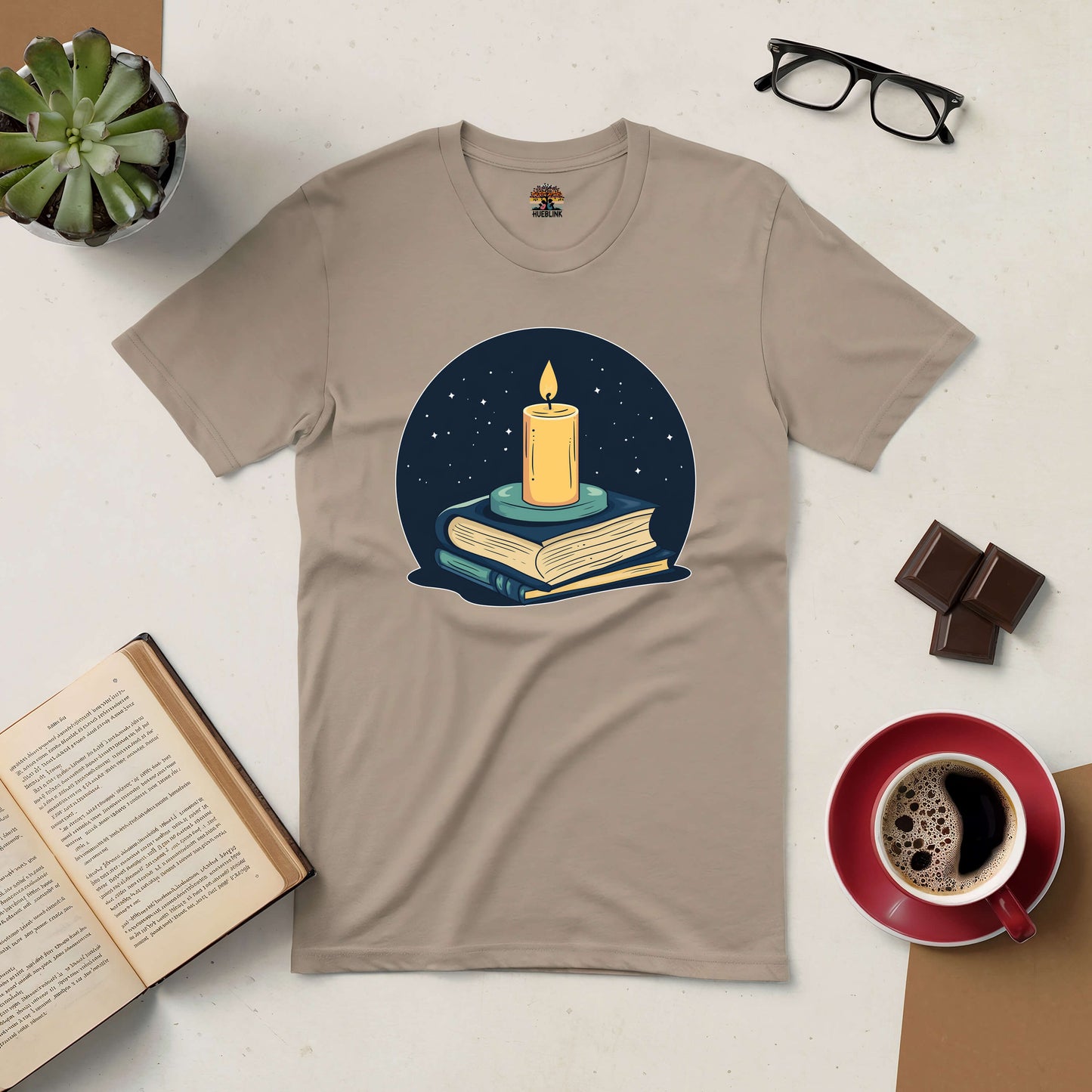 "Illuminate Your Mind Tee with candle and books design, surrounded by coffee, glasses, chocolate, and an open book."