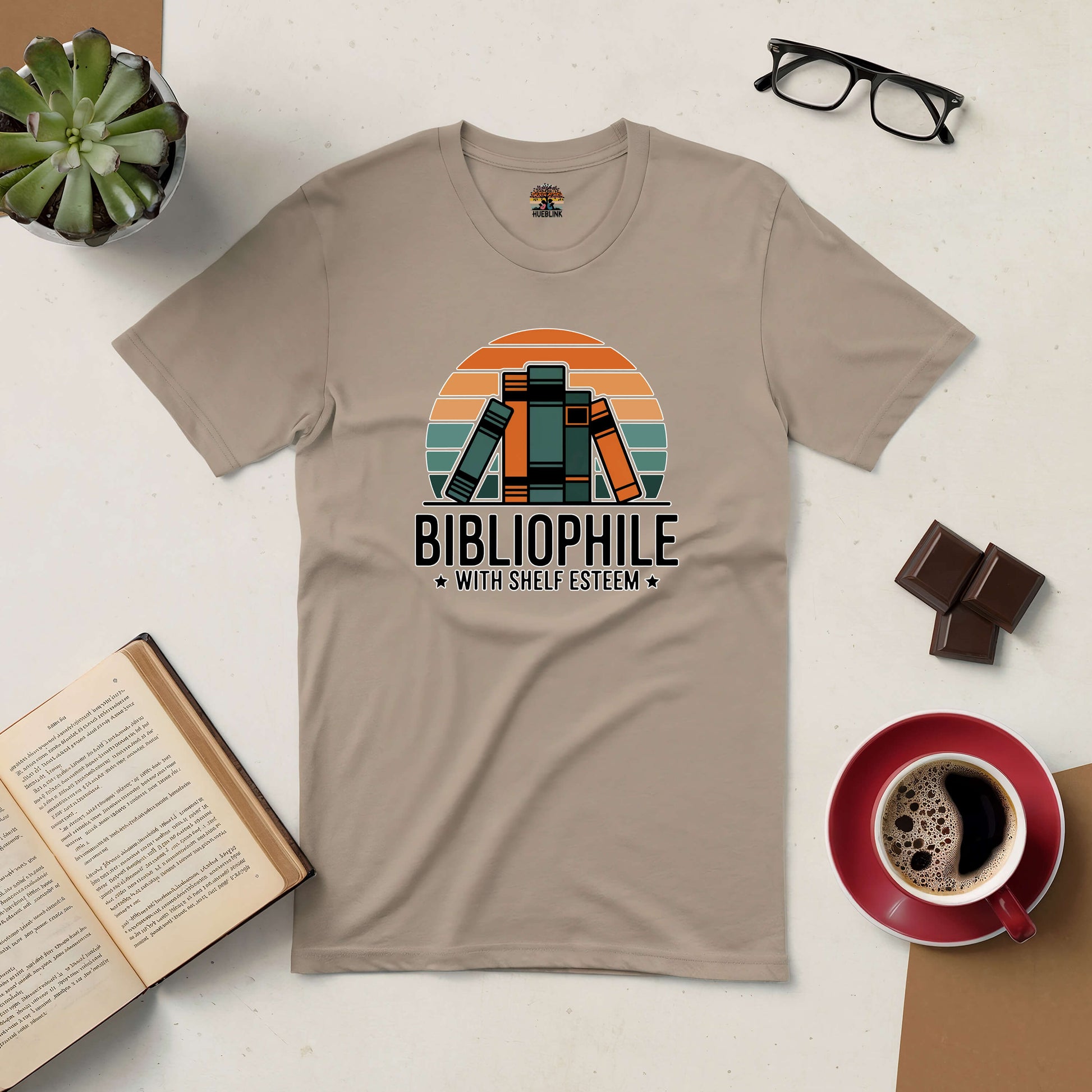 Beige retro-style "Bibliophile with Shelf Esteem" tee on a flatlay with coffee, book, glasses, and plant for book lovers.