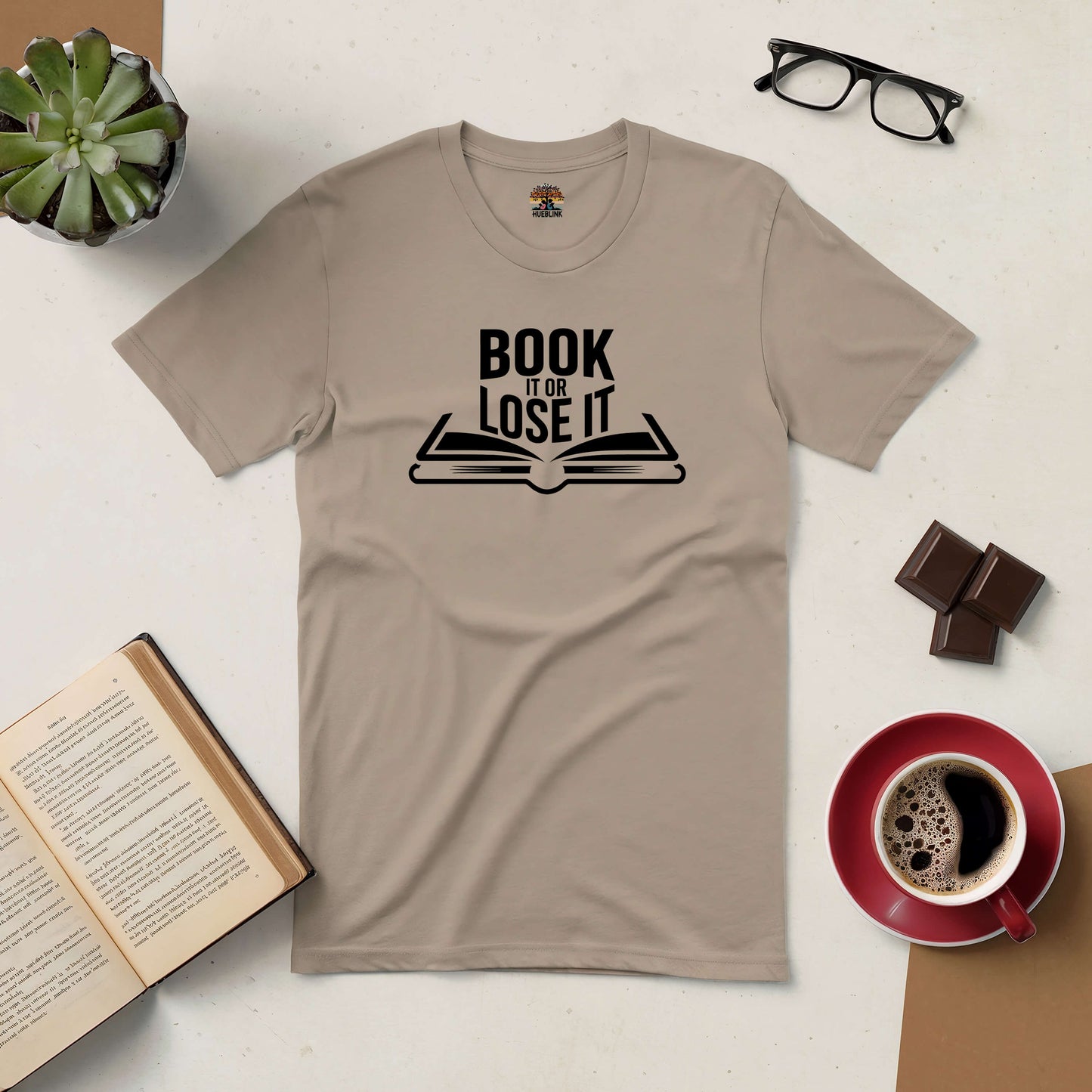 "Beige Book It or Lose It Tee with reading motif, surrounded by book, coffee, glasses, and plants"