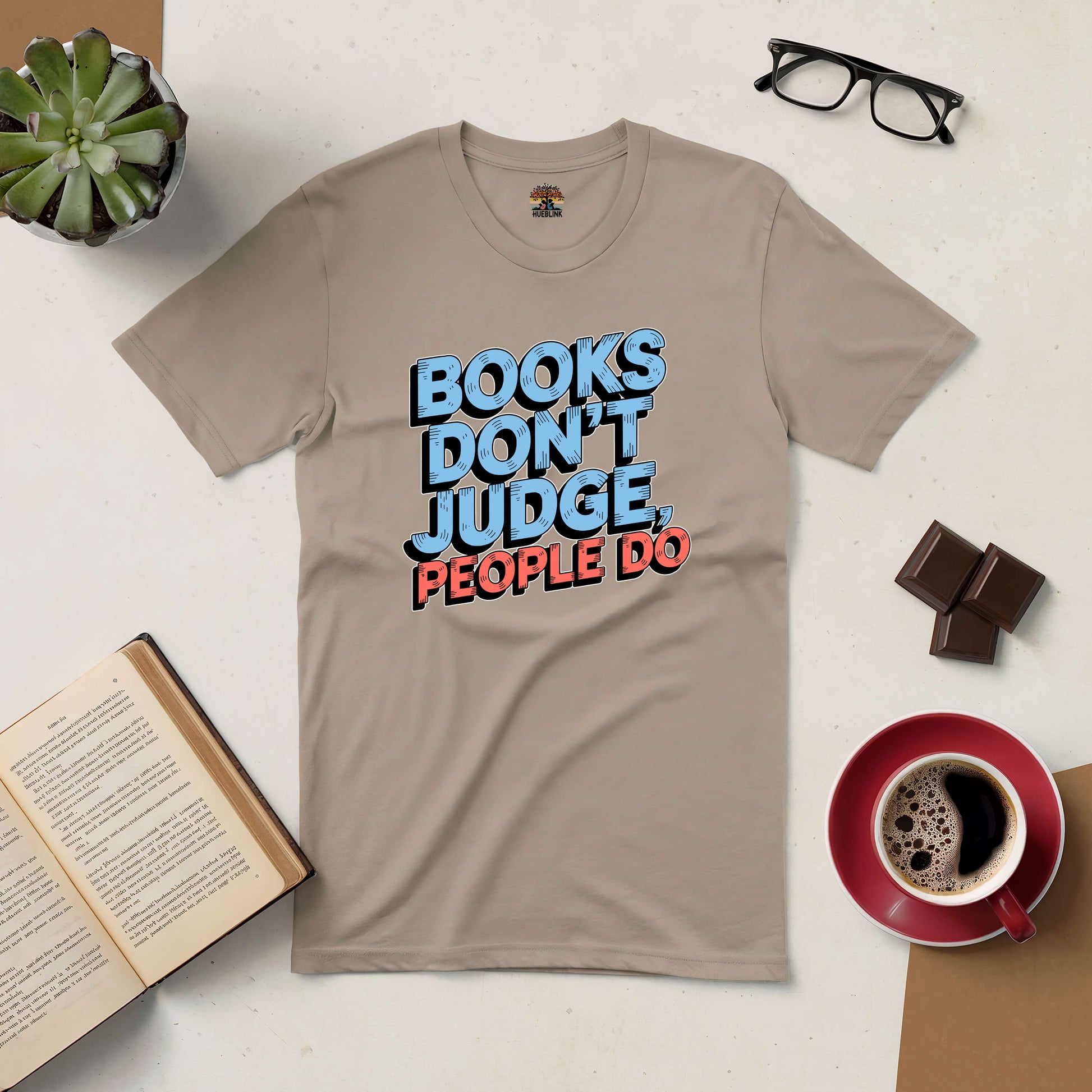 Beige tee with "Books Don't Judge, People Do" text, surrounded by an open book, coffee, glasses, and chocolate on a table.