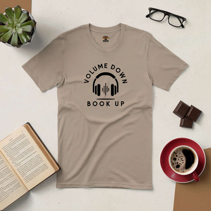 Beige "Volume Down, Book Up" tee with headphones design, surrounded by a book, coffee mug, eyeglasses, and chocolate pieces.