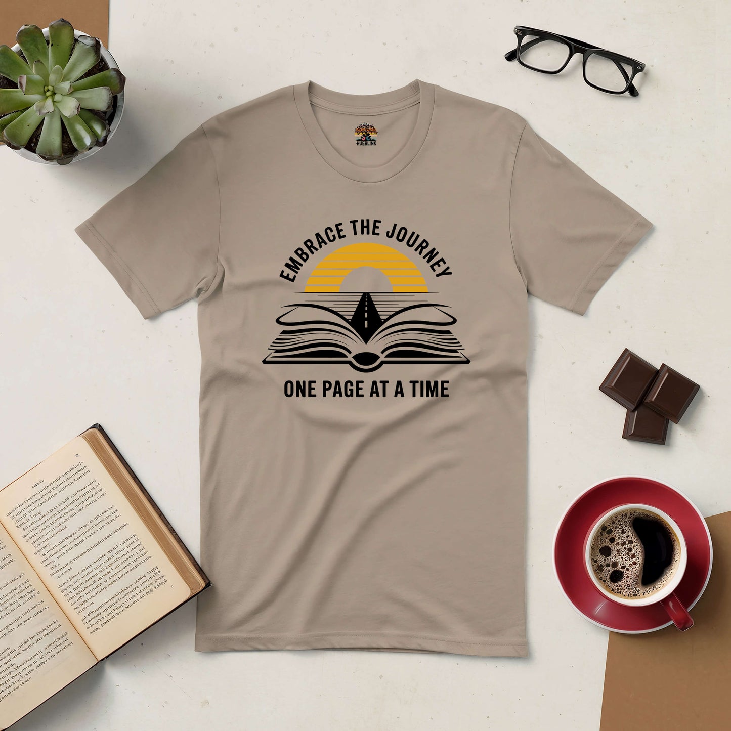 Beige "Embrace the Journey" tee with open book and road design, surrounded by glasses, coffee, chocolate, and a succulent.