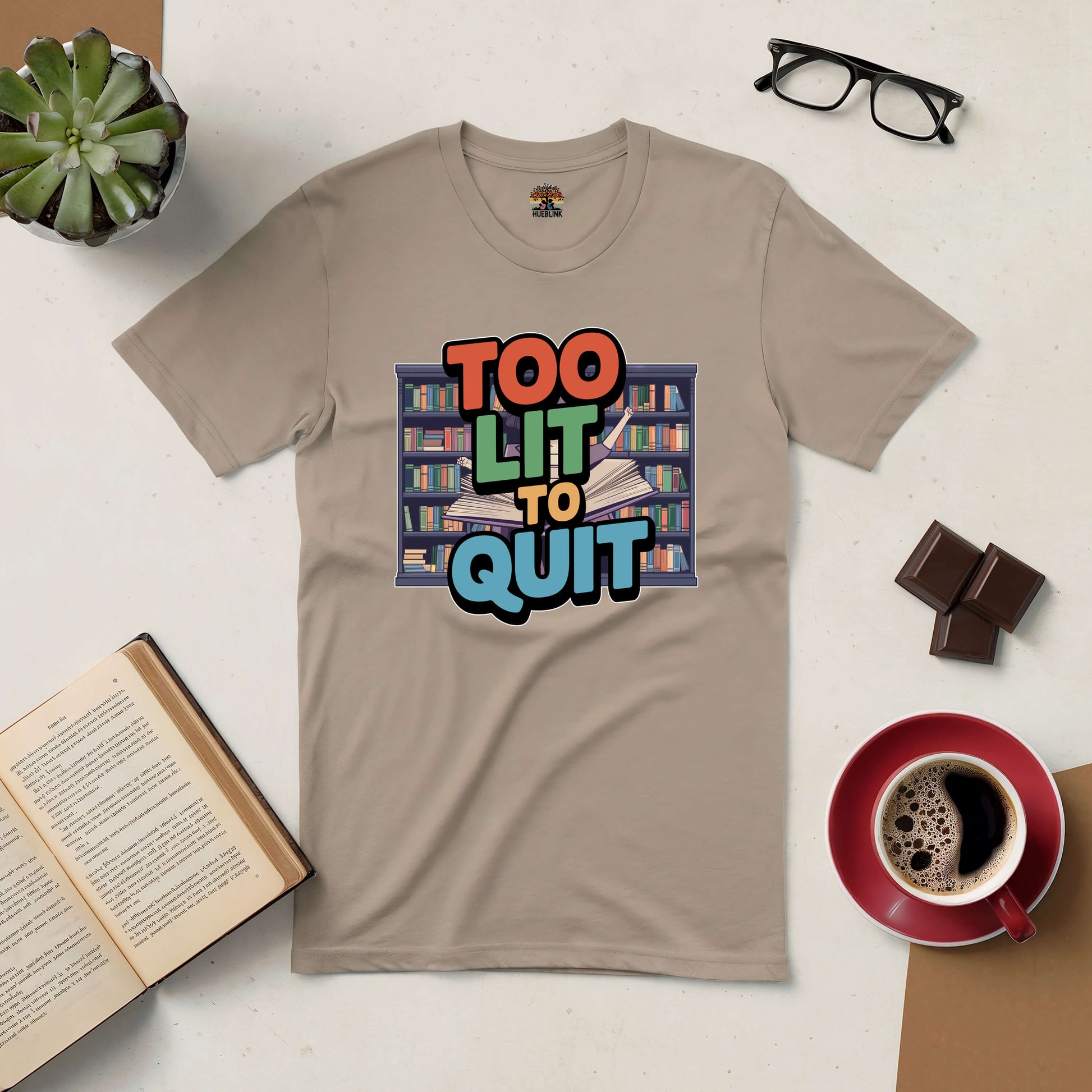 Beige "Too Lit To Quit" tee with book graphic, surrounded by open book, coffee, glasses, chocolate, and a succulent. Perfect for book lovers.