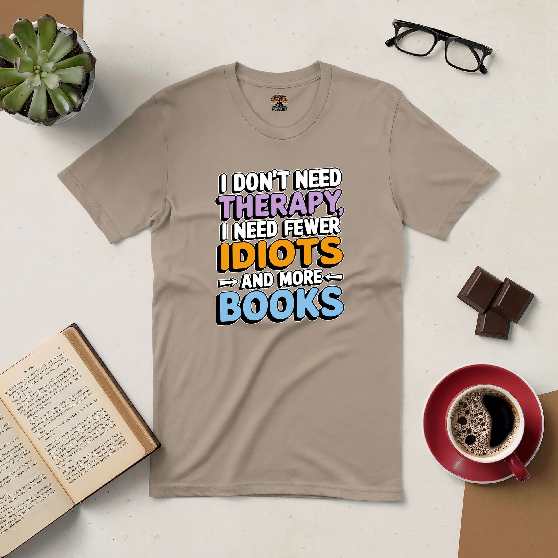 Beige tee with "I Don't Need Therapy, I Need Fewer Idiots and More Books" text, displayed with a book, coffee, and glasses.