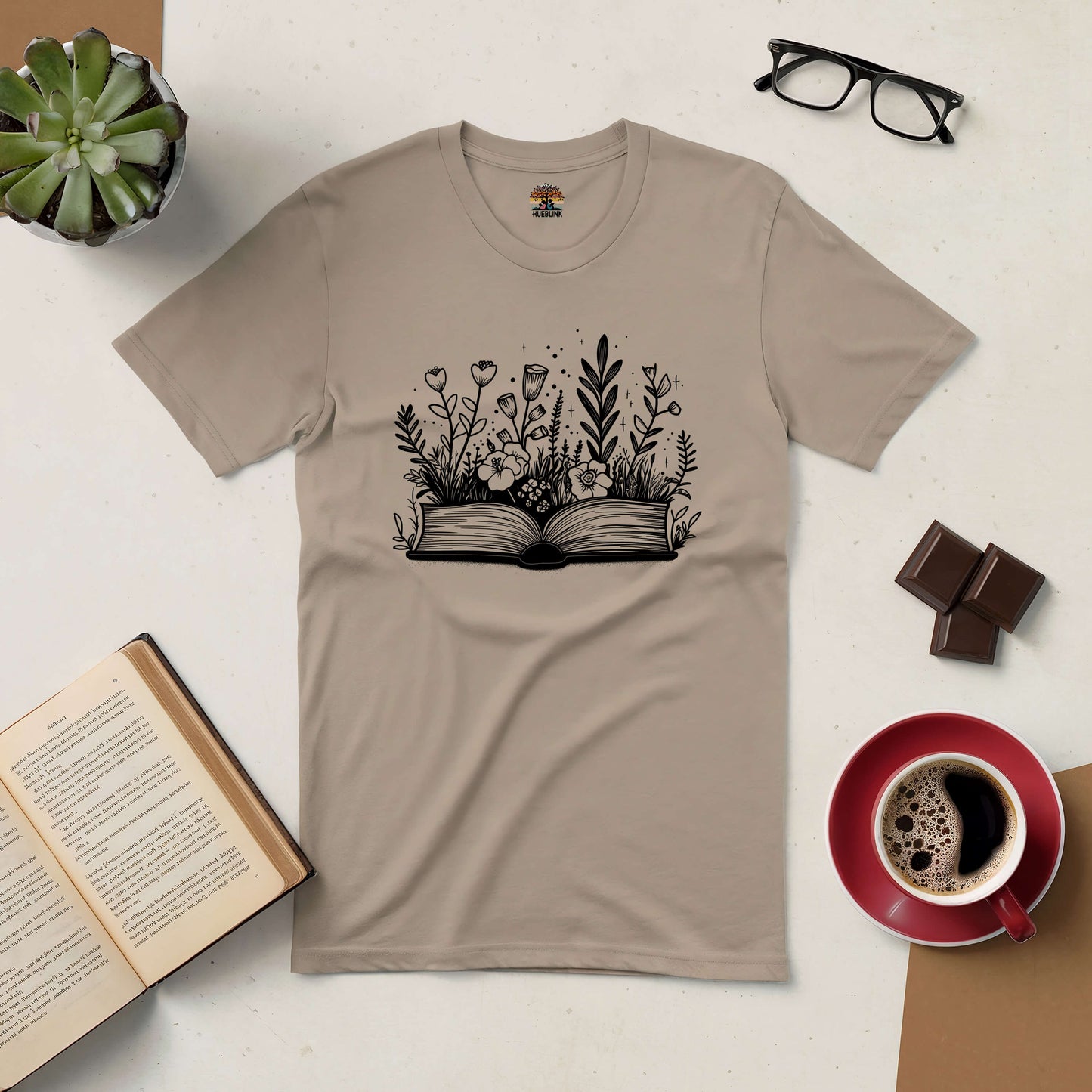 Books and Blooms tee with open book and floral design, surrounded by coffee, chocolate, glasses, and a succulent plant.