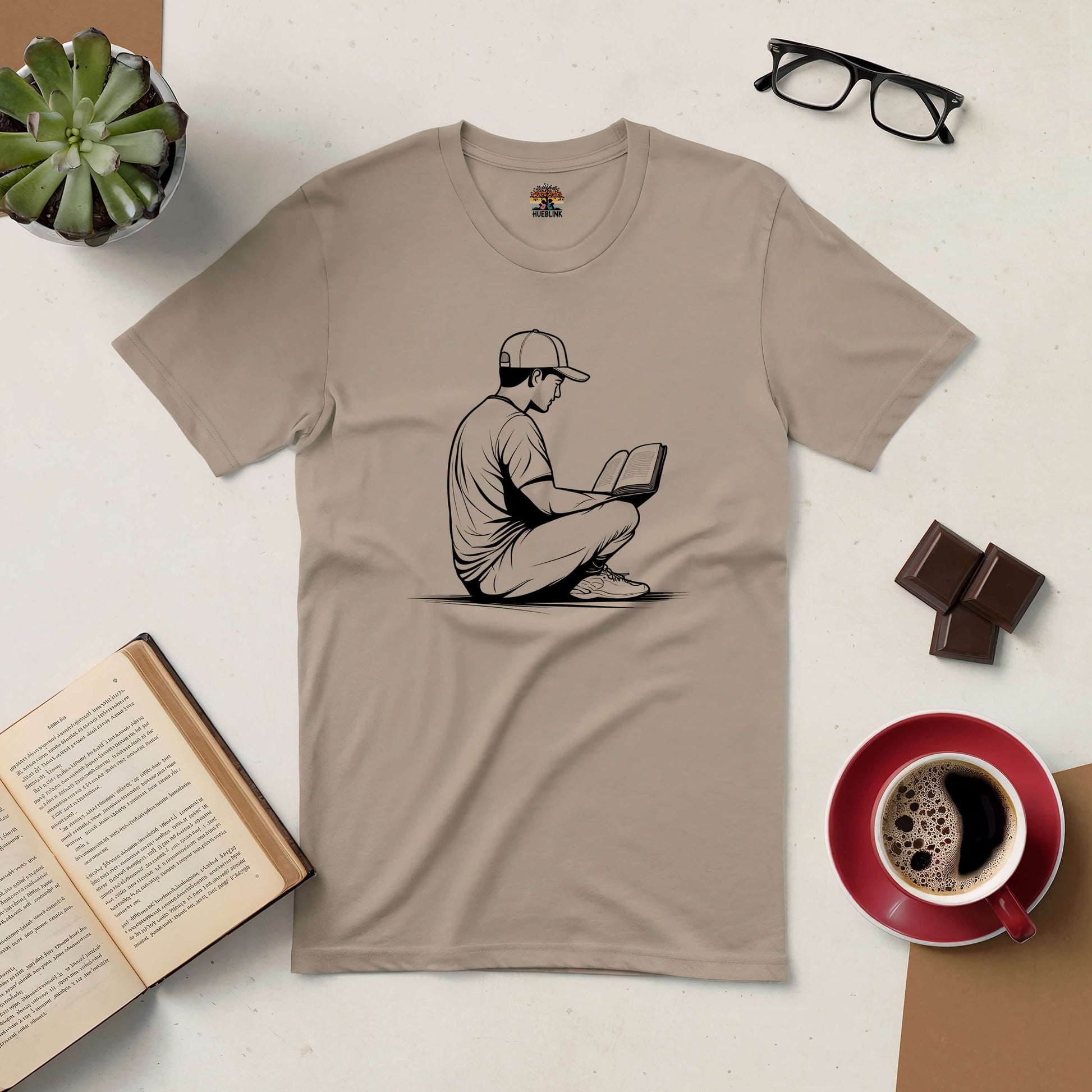 Beige T-shirt with reading figure design, surrounded by a book, glasses, coffee, chocolate, and a plant on a white surface.