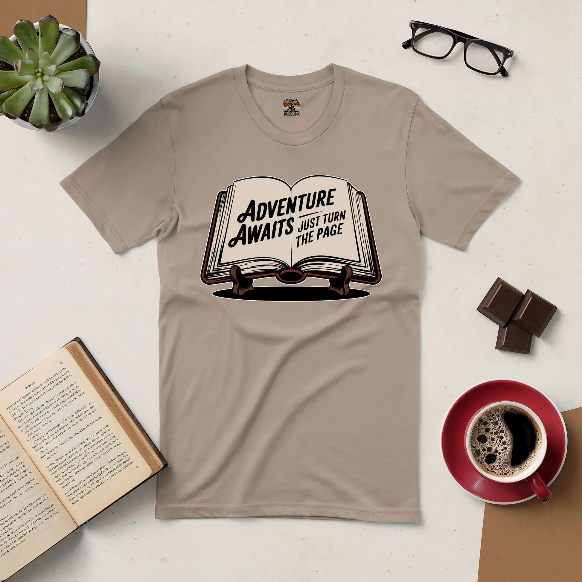 Beige Adventure Awaits tee with book graphic and 'Just Turn the Page' text, surrounded by coffee, chocolate, book, and glasses.