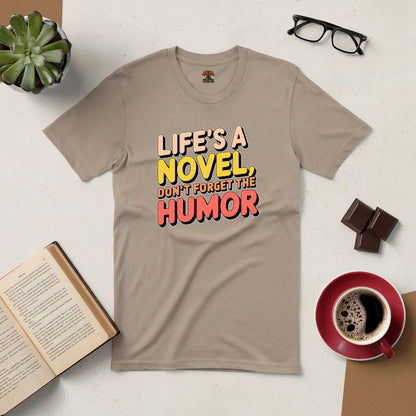 Beige tee with "Life's a Novel, Don't Forget the Humor" text, surrounded by books, glasses, plant, and coffee. Perfect for witty readers.