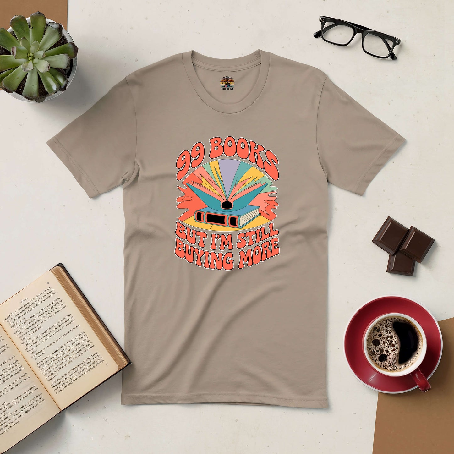 Beige "99 Books and Counting!" tee with colorful design, surrounded by coffee, open book, and glasses, showcasing a reader's passion.