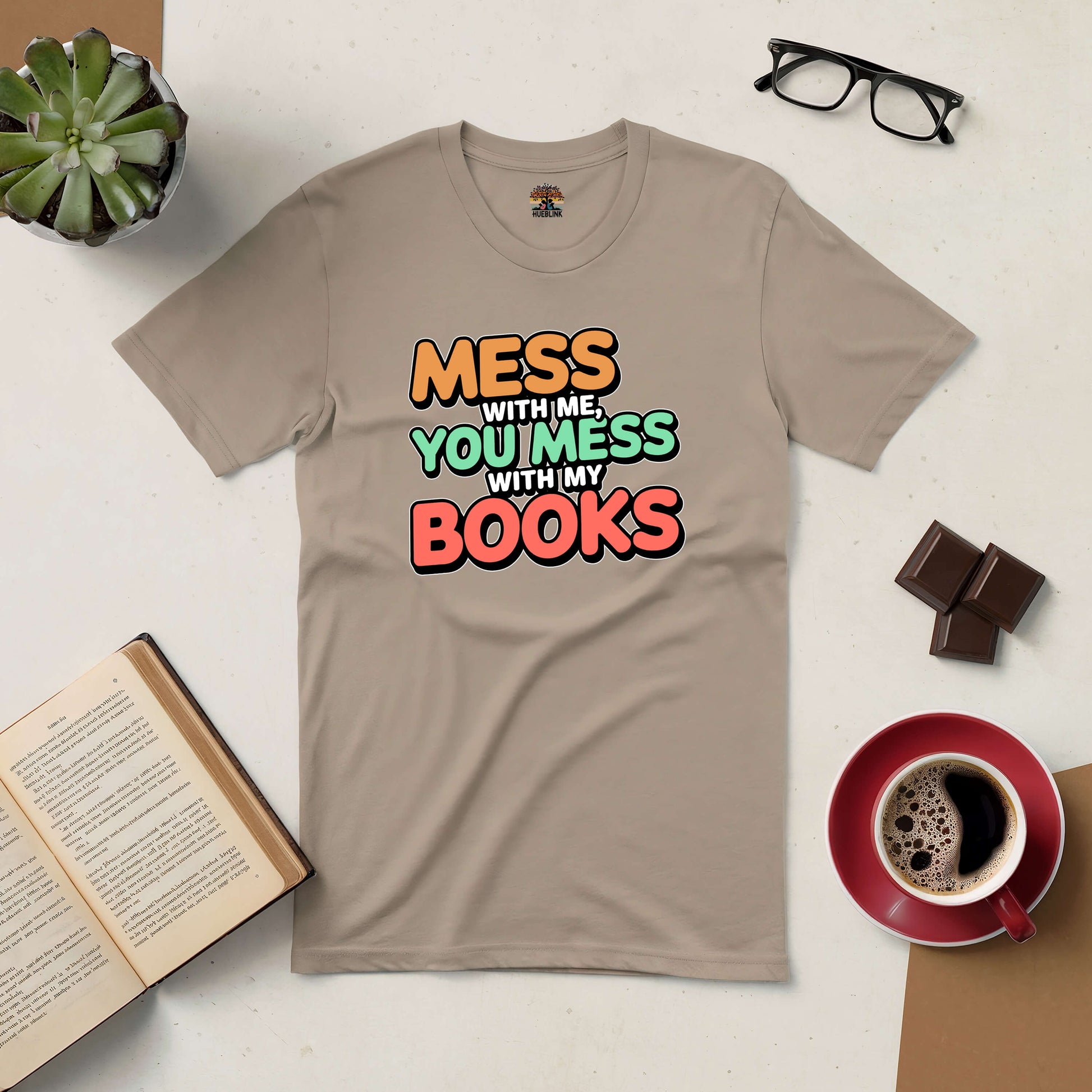 Tan t-shirt with "Mess With Me, You Mess With My Books" slogan, surrounded by coffee, chocolate, glasses, and an open book.