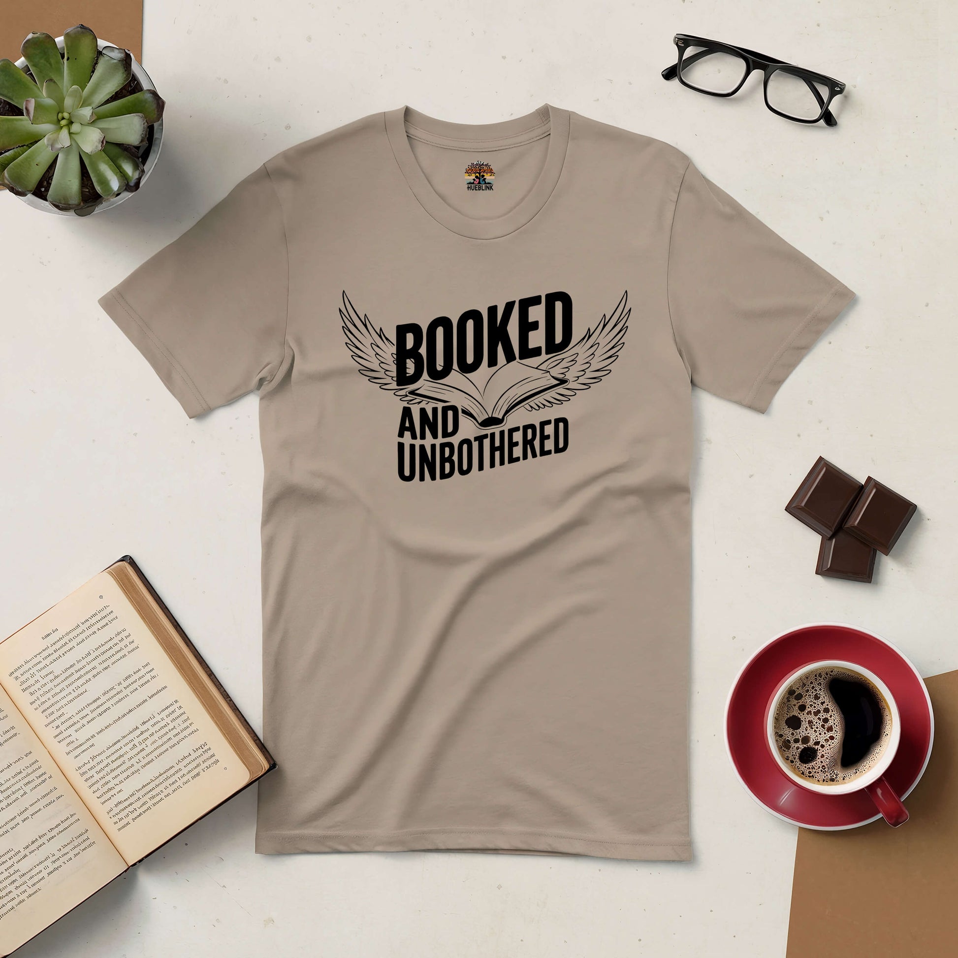 "Booked and Unbothered Tee for avid readers, with open book graphic, surrounded by glasses, coffee, chocolate, plant, and open book"