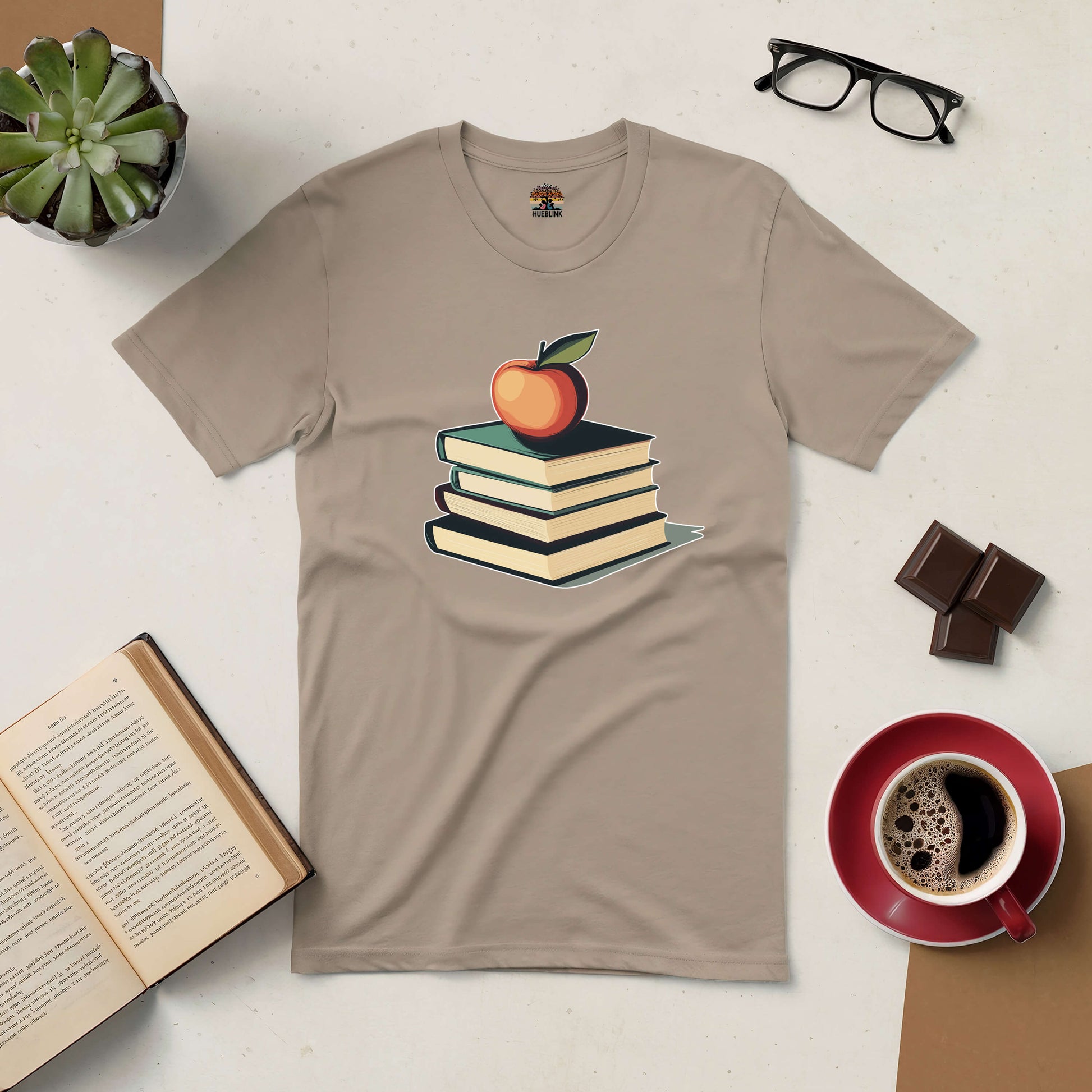 Beige tee with stacked books and apple design, for book lovers and knowledge seekers. Coffee, glasses, and chocolate on table.