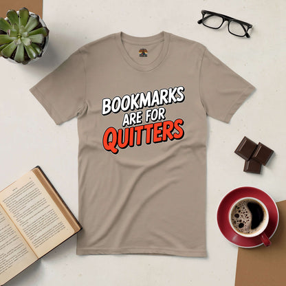 Beige tee with bold text "Bookmarks Are for Quitters," surrounded by glasses, chocolate, coffee, a book, and a potted plant.