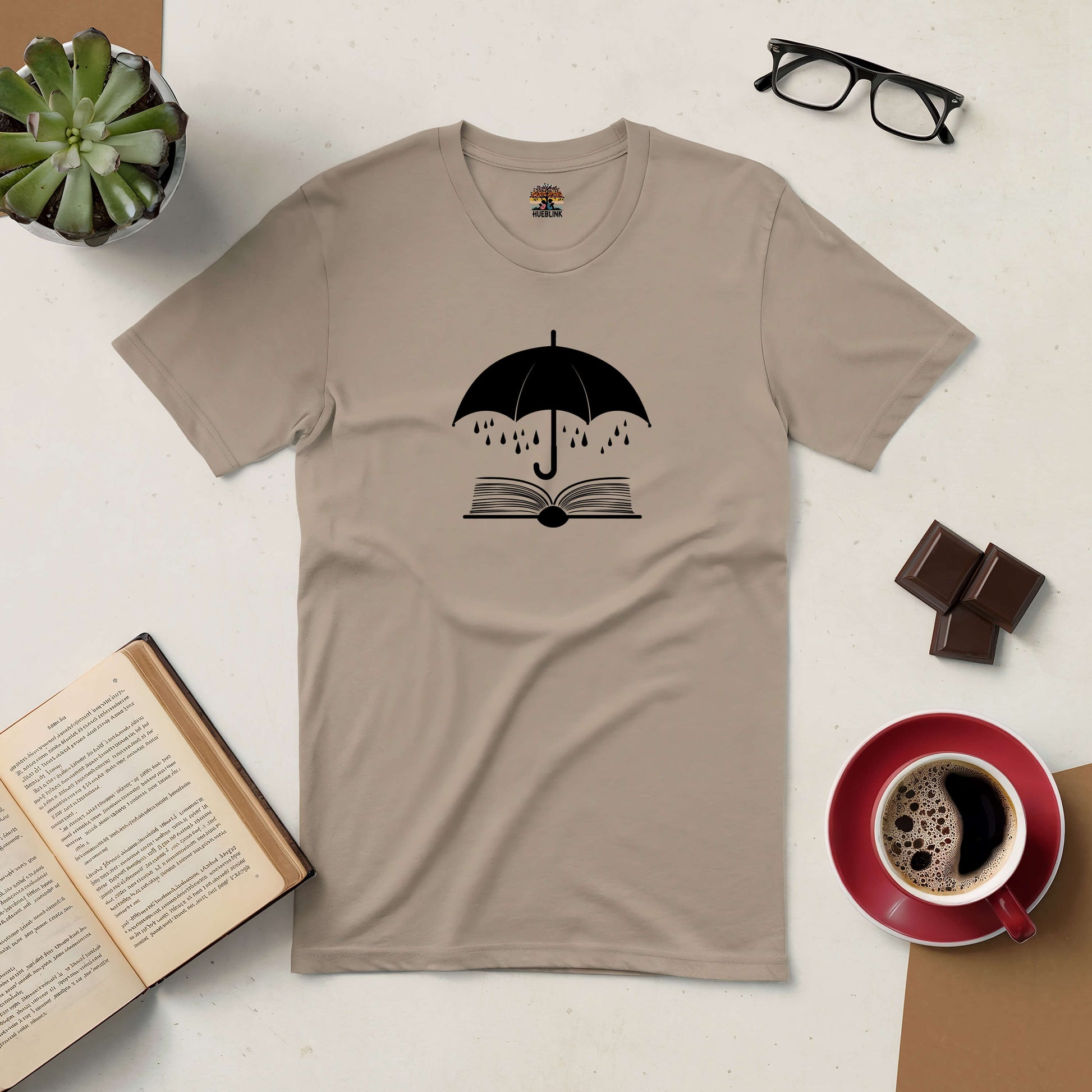 Sheltering Stories Tee with book and umbrella design, surrounded by coffee, chocolate, open book, plant, and glasses.