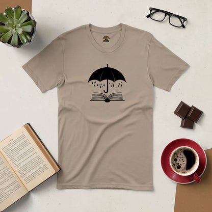 Sheltering Stories Tee with book and umbrella design, surrounded by coffee, chocolate, open book, plant, and glasses.