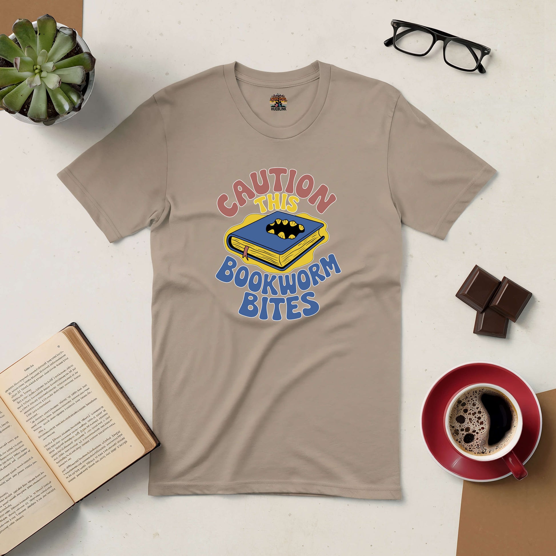 "Caution, This Bookworm Bites Tee with playful book design surrounded by coffee, chocolates, and glasses on a table"