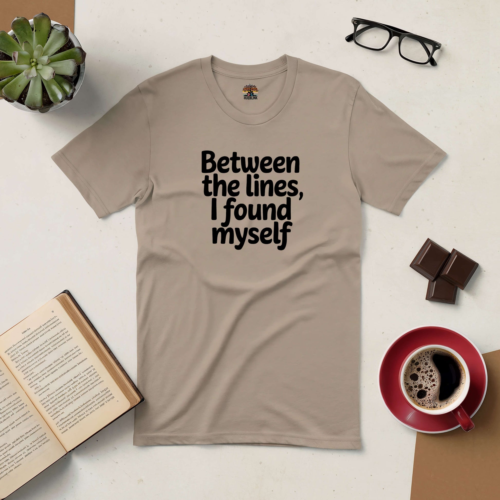 Beige "Between The Lines, I Found Myself" tee styled with a book, coffee cup, chocolates, glasses, and a plant. Perfect for introspective readers.