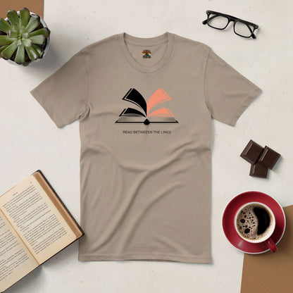 Beige tee with "Read Between The Lines" design, surrounded by coffee, glasses, chocolate, and an open book on a white table.