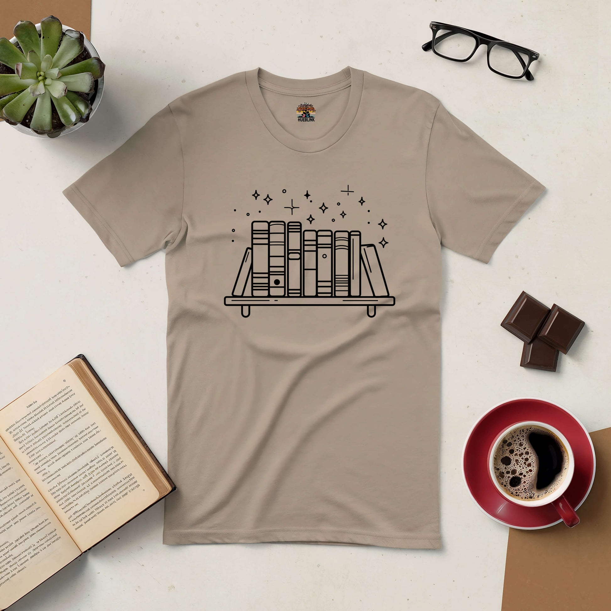 Beige t-shirt with bookshelf and sparkles design, surrounded by coffee, chocolate, glasses, book, and plant.