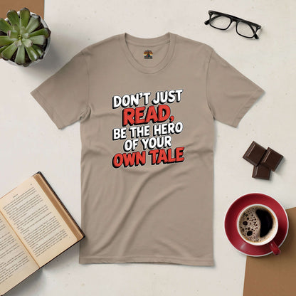 "Be the Hero of Your Own Tale tee featuring motivational text, surrounded by book, coffee, and glasses on a desk."