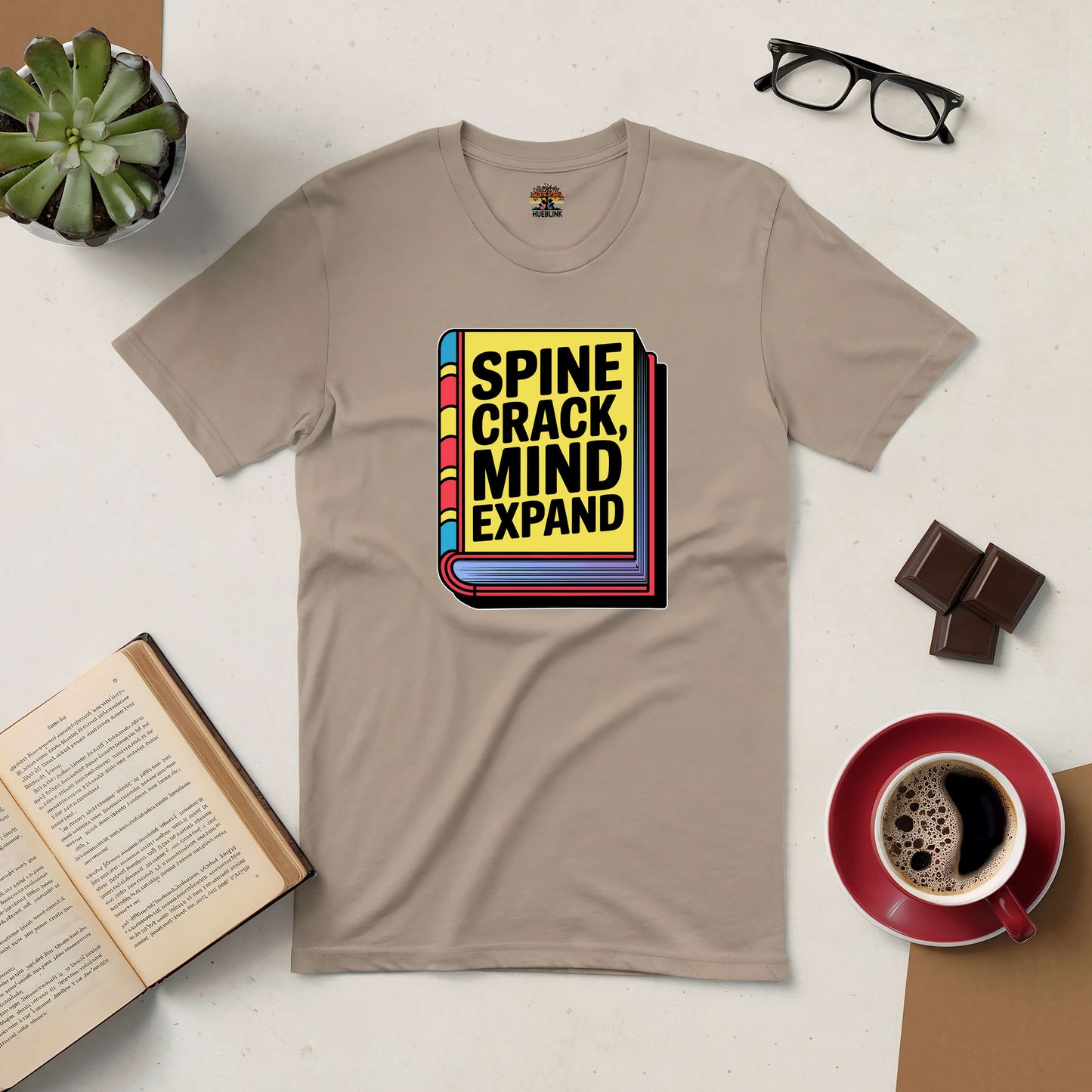 Spine Crack, Mind Expand Tee with book graphic, surrounded by coffee, glasses, and chocolate on a light surface.